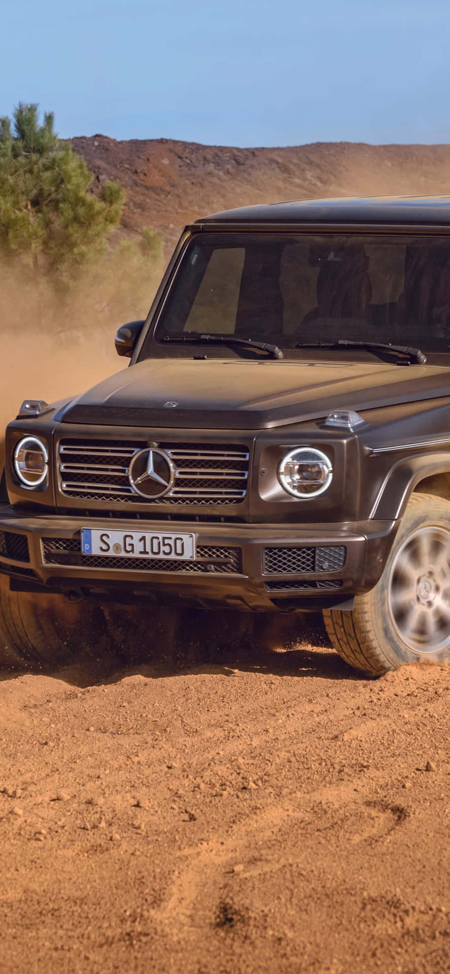 黑色 Mercedes G-Class 在塵土飛揚的沙漠地形中行駛，有丘陵和樹木。