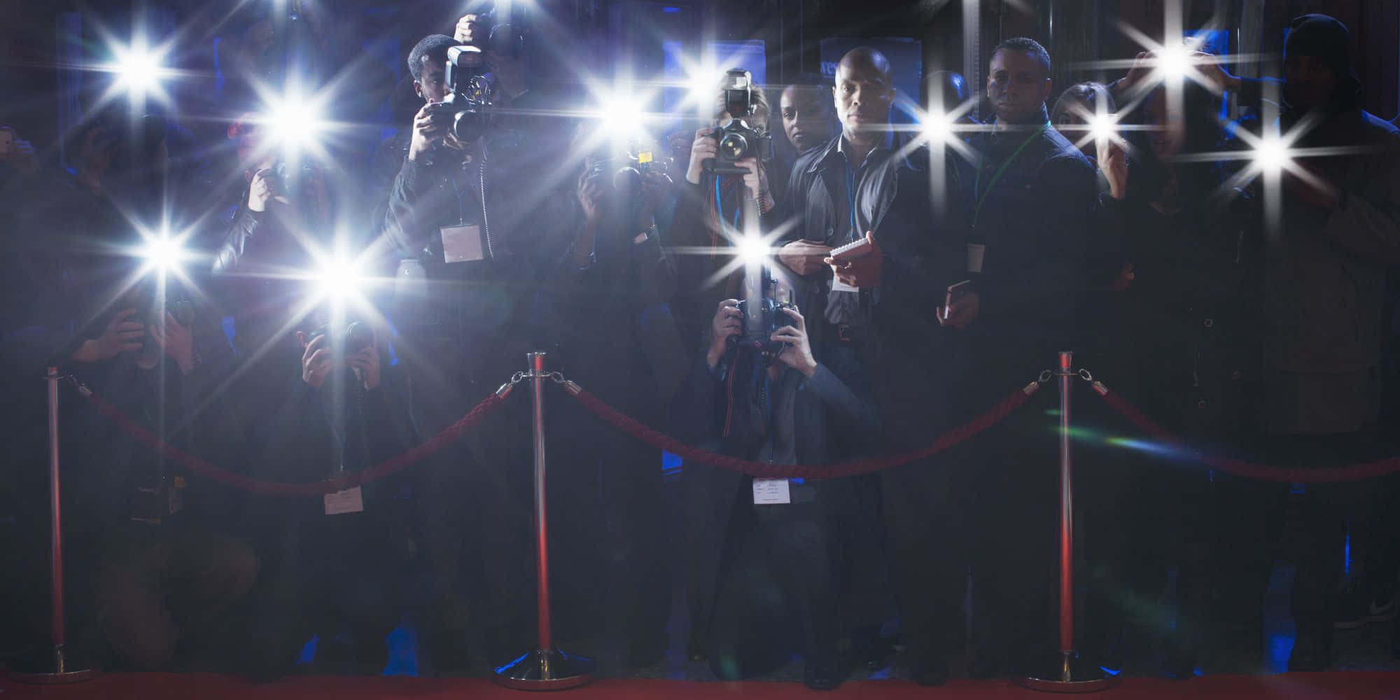 Paparazzi 桌布 壁紙 — 攝影師們帶著閃光燈的相機，在明亮的攝影棚燈光下，擠在紅地毯的天鵝絨繩索後面。