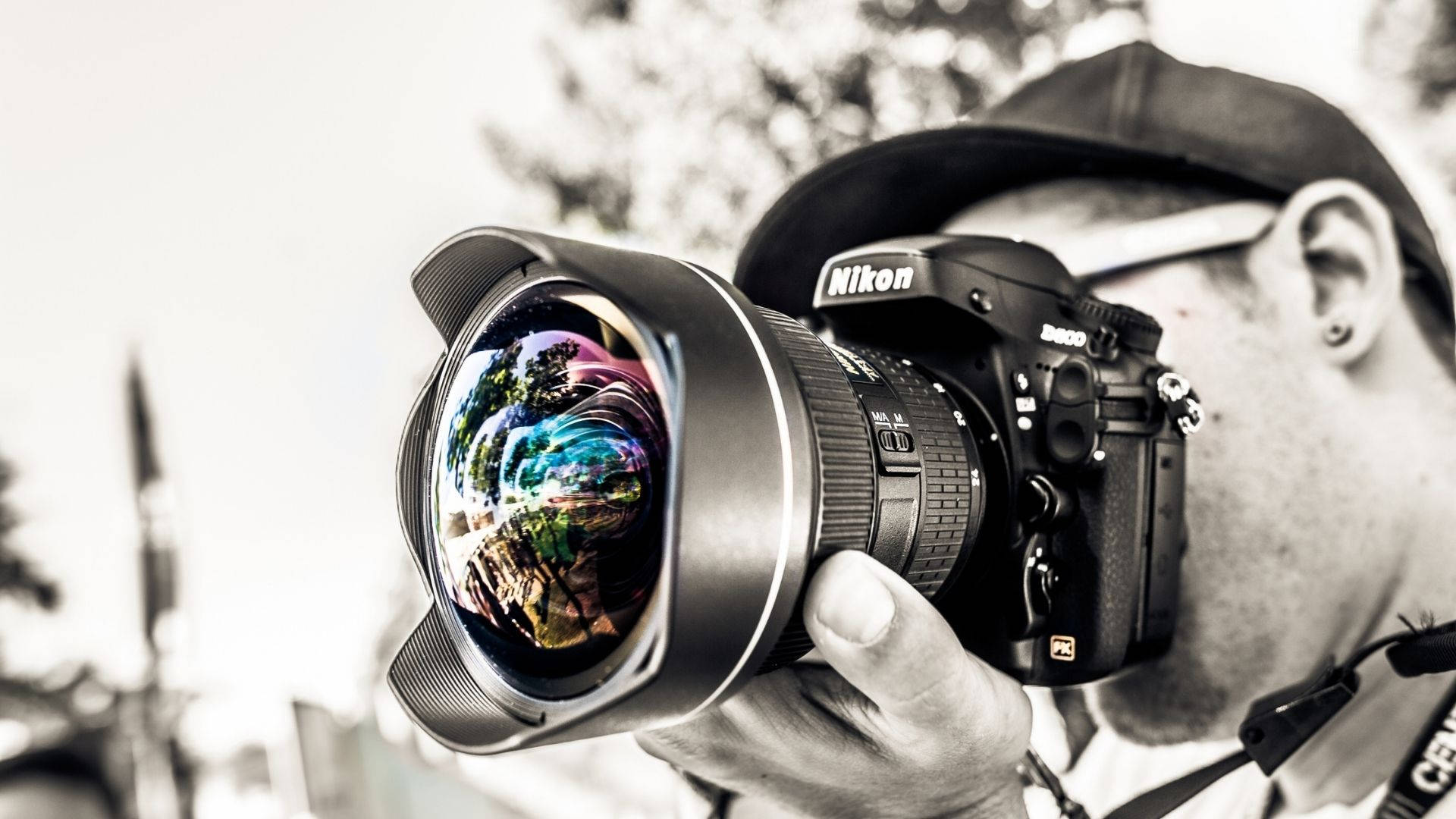 攝影師彩虹鏡頭 壁紙 — 一位攝影師手持一部Nikon DSLR相機，鏡頭上有鮮豔的彩虹反射，背景模糊。