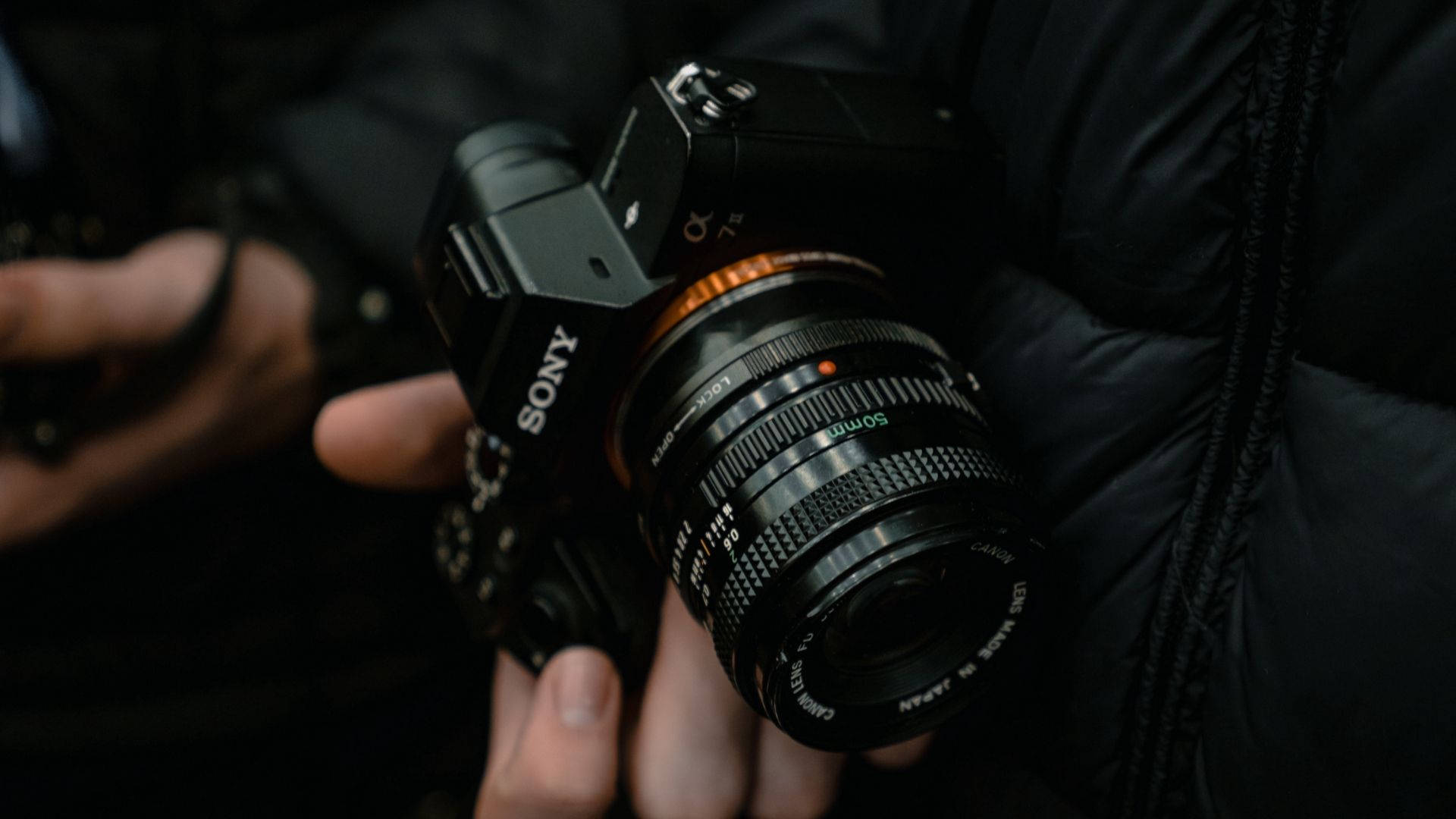 攝影師手持經典相機 壁紙 — 黑色Sony無反相機配Canon鏡頭，手持於深色羽絨外套背景前的特寫。