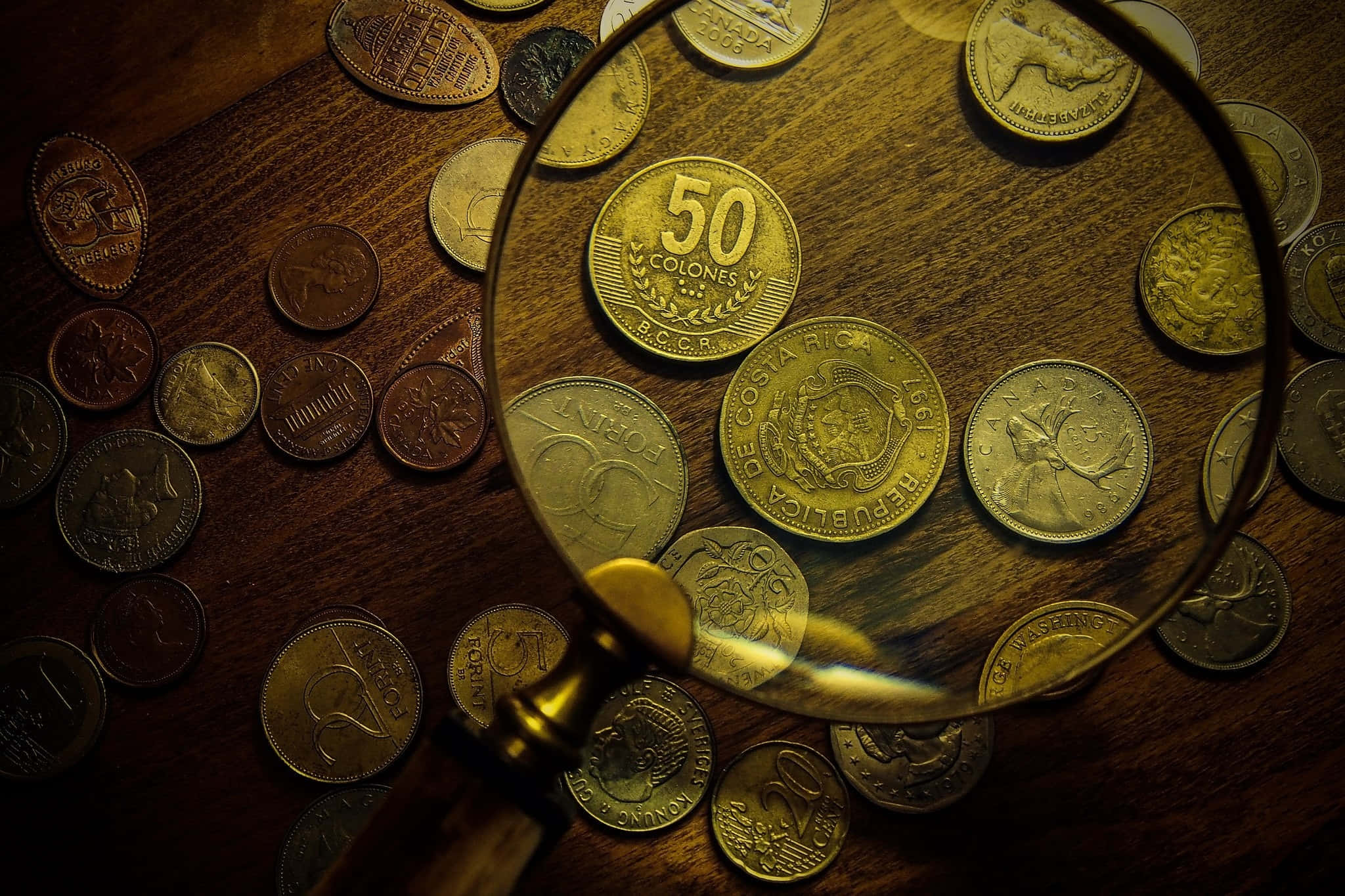 放大鏡與 50 美分硬幣 壁紙 — 放大鏡懸浮在散落的哥斯達黎加硬幣上方，置於深色木桌上，溫暖燈光下。