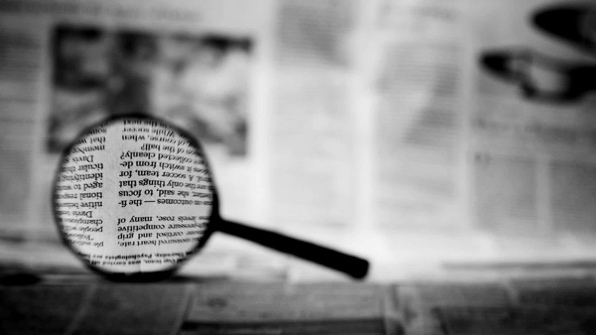放大鏡與調查新聞的神秘感 壁紙 — 放大鏡清晰地聚焦在文字上，而周圍的報紙則模糊成柔和的灰階色調。