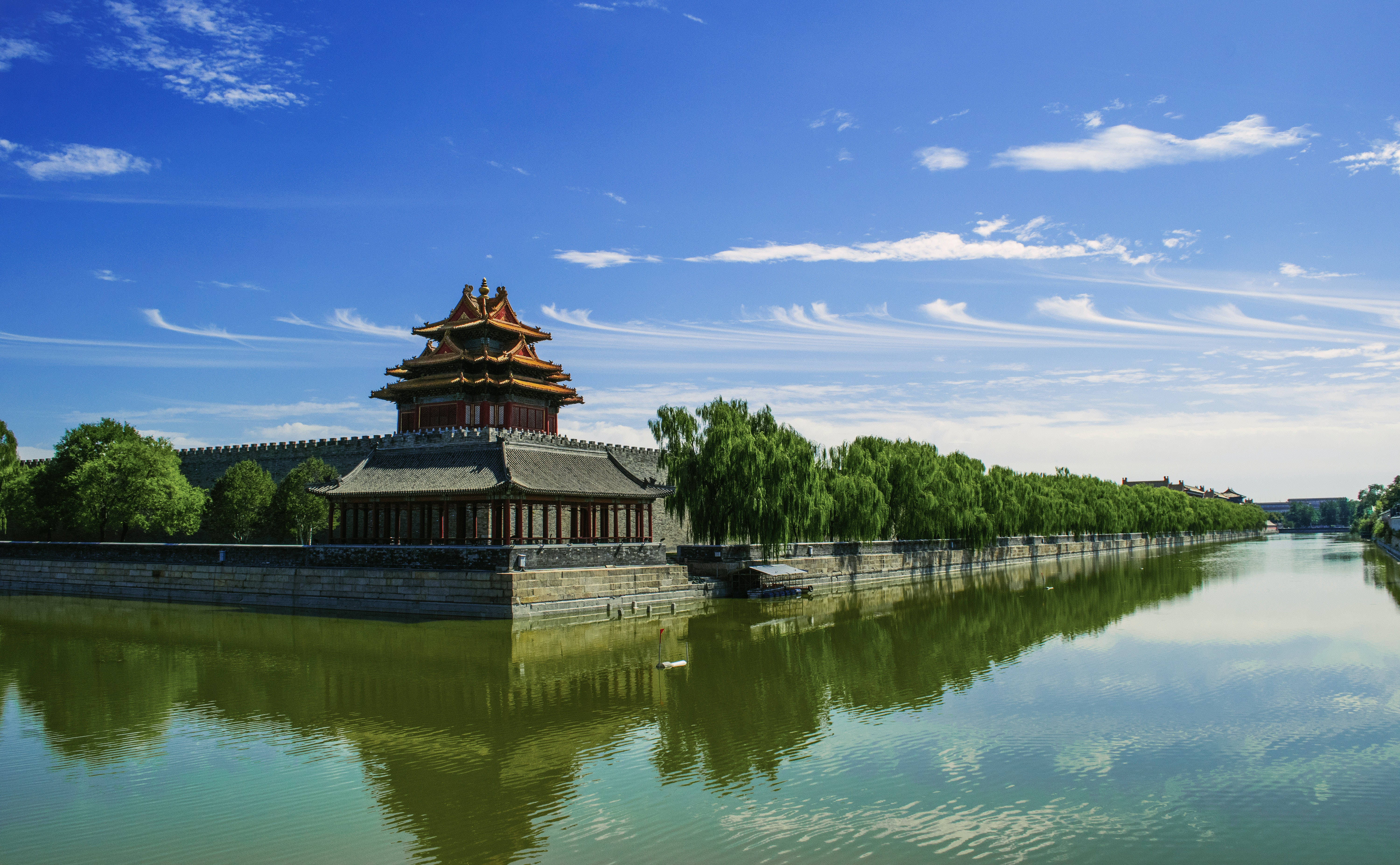 故宮博物院河景 壁紙 — 一座傳統中式宮殿塔樓，紅瓦灰磚，雄偉地矗立在平靜的護城河旁，倒映著藍天和綠樹。