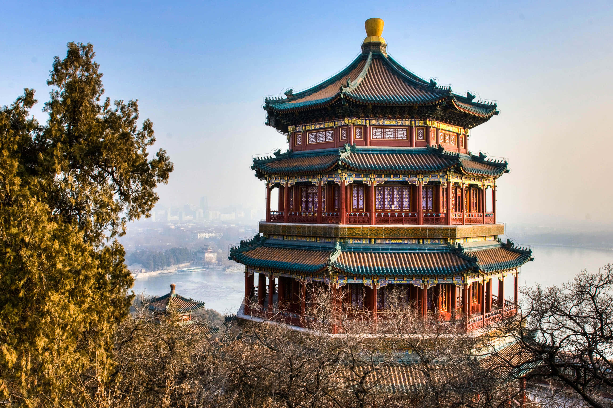 故宮壯麗的塔樓 壁紙 — 一座雄偉的傳統中式寶塔，紅色柱子和綠色瓦片屋頂，矗立在山丘上，俯瞰著寧靜的湖泊和遠處的城市景觀，天空清澈湛藍。