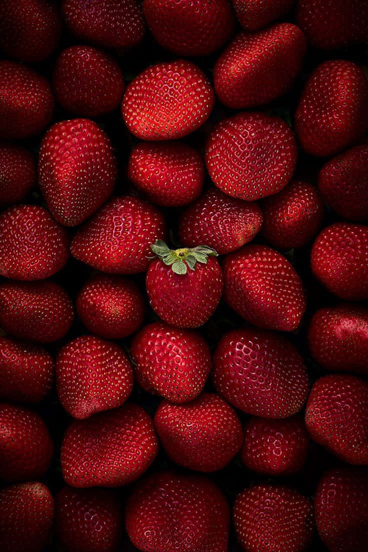 散發熱烈氣息 壁紙 — 一簇密集的成熟紅色草莓，其中一顆帶綠色頂部的莓果在深色背景上居中。