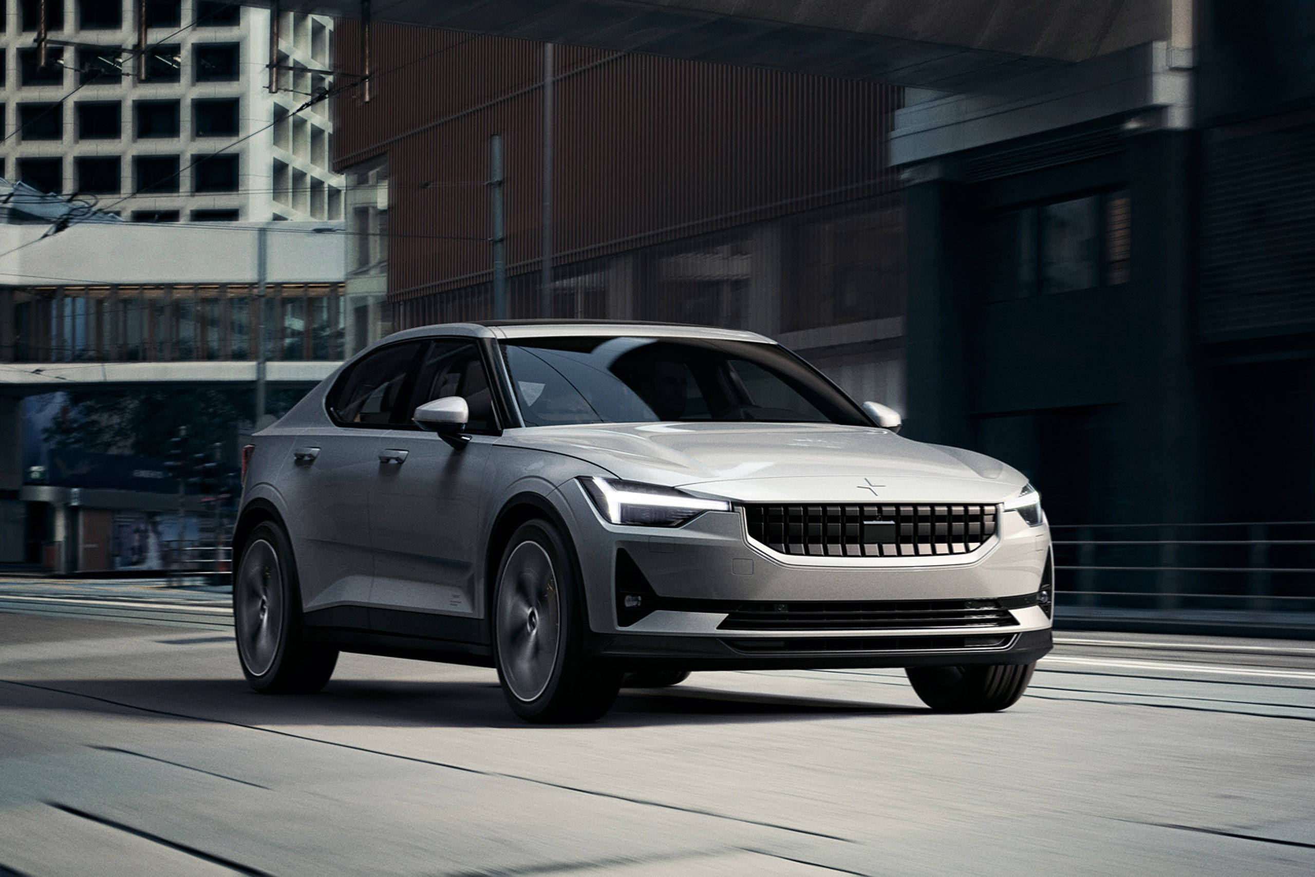 數位渲染Polestar 2 壁紙 — 一輛時尚的銀色電動車在明亮的日光下滑過一條擁有玻璃和鋼鐵建築的現代城市街道。