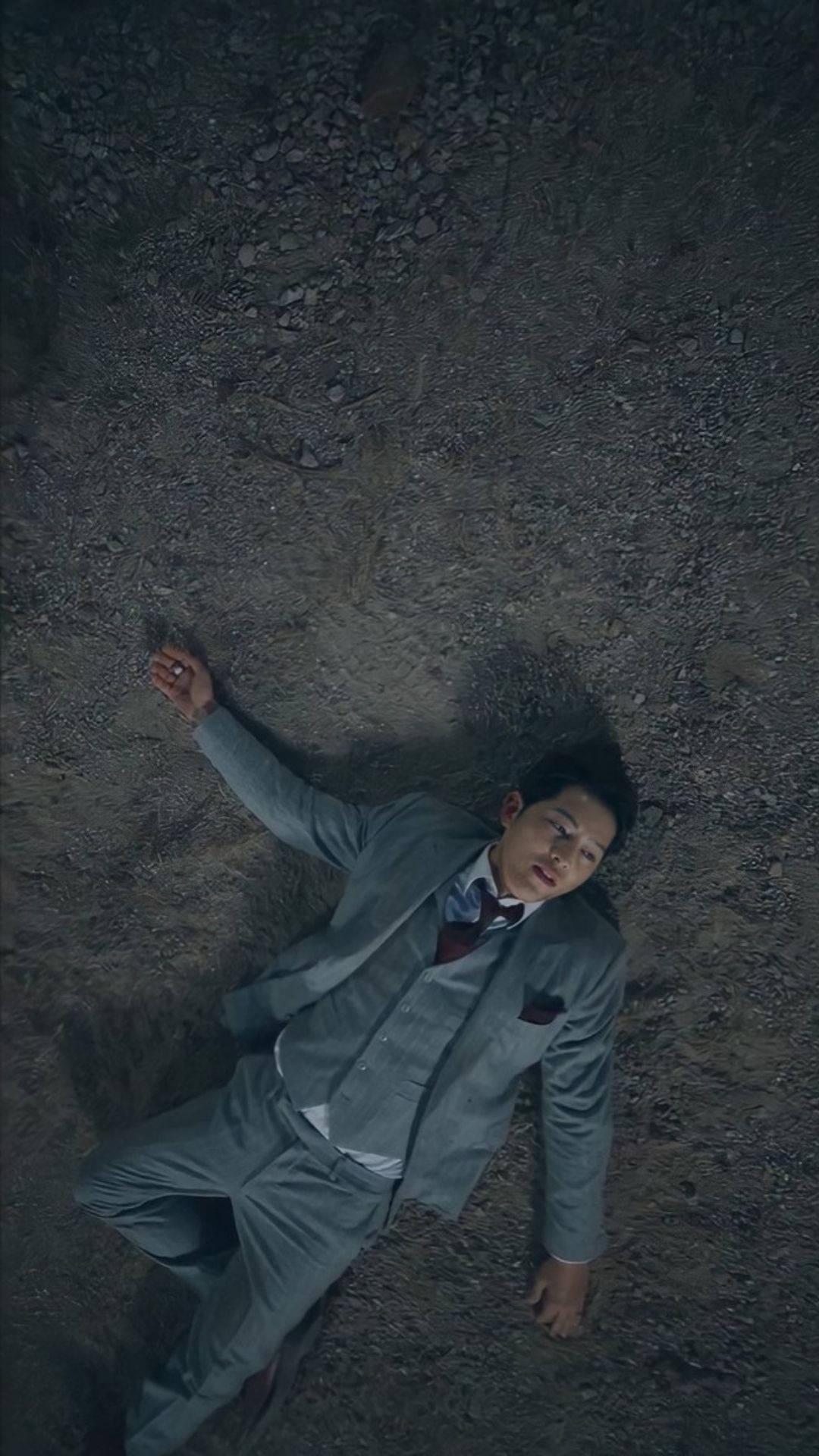 文森佐臥地 壁紙 — 一名身穿灰色西裝的男子在昏暗、陰鬱的燈光下一動不動地躺在碎石上。