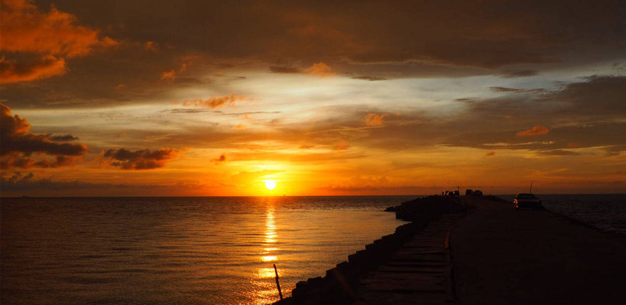 文萊橙色夕陽天空 壁紙 — 一輛汽車在日落時沿著碼頭行駛，金色光線映照在平靜的水面上，天空佈滿戲劇性的橙色雲彩。