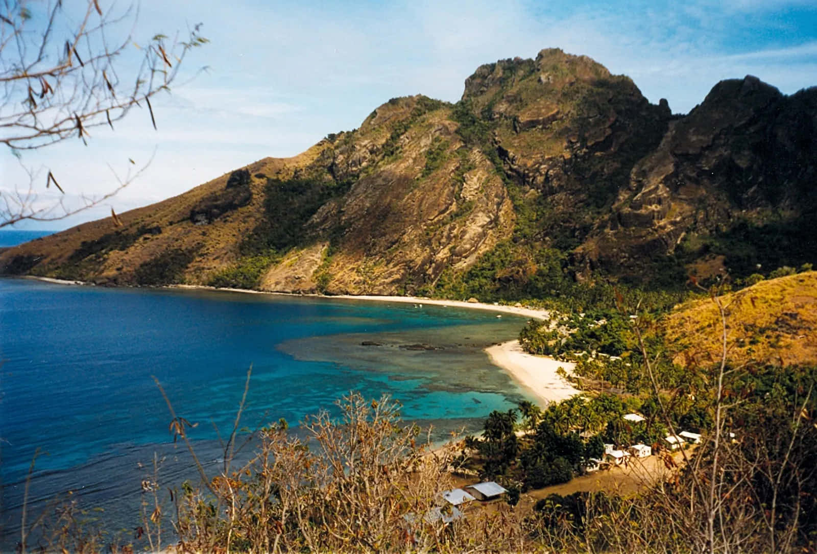 斐濟島壯麗全景 壁紙 — 一個隱蔽的熱帶海灣，碧綠的海水環繞著白色沙灘，沙灘坐落在崎嶇的綠色山脈之間，上方是清澈的藍天。