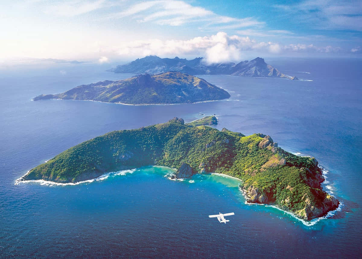 斐濟島壯麗景色 壁紙 — 從空中俯瞰鬱鬱蔥蔥的綠色島嶼，擁有碧綠色的潟湖，被深藍色的海洋環繞，天空部分多雲。