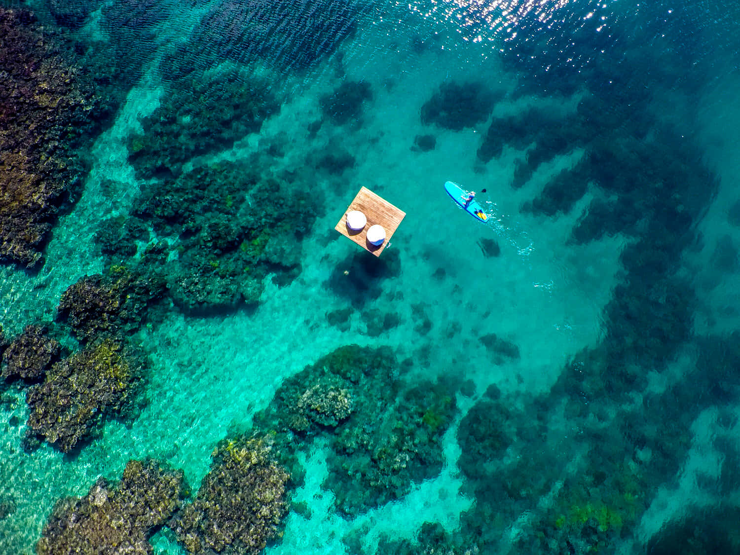 斐濟島漂浮竹筏圖 壁紙 — 從空中俯瞰，一名槳板衝浪者滑過一個木製平台，平台上有兩把白色遮陽傘，海水清澈碧藍。