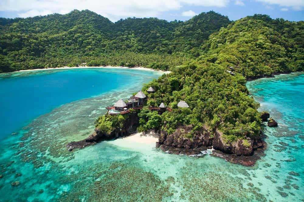 斐濟島隱藏小屋度假村 壁紙 — 鳥瞰一個鬱鬱蔥蔥的熱帶島嶼，擁有碧綠的海水、珊瑚礁和坐落在白色沙灘上的茅草屋頂小屋。