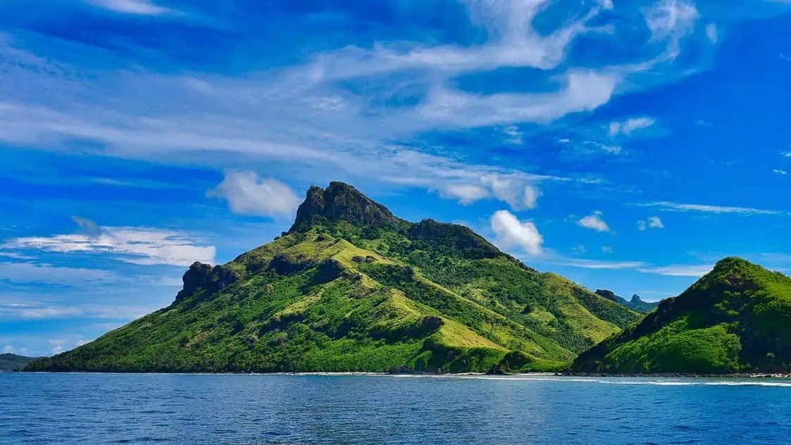 斐濟島高聳綠山景圖 壁紙 — 一個鬱鬱蔥蔥的綠色火山島從平靜的藍色海水中陡峭升起，上方是充滿活力的藍天和縷縷白雲。