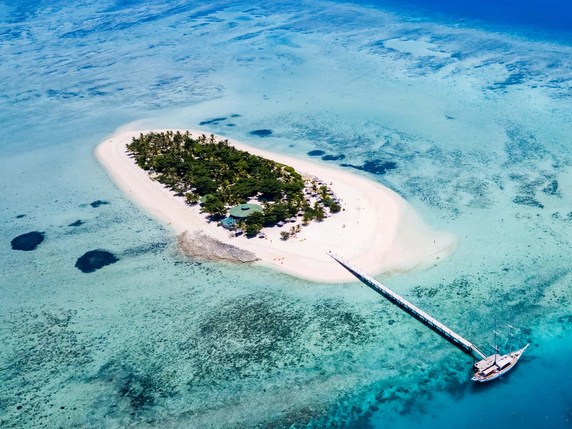 斐濟群島令人屏息的美景 壁紙 — 熱帶島嶼的鳥瞰圖，有白色沙灘棕櫚樹和停泊在長碼頭的船隻，海水清澈碧綠。