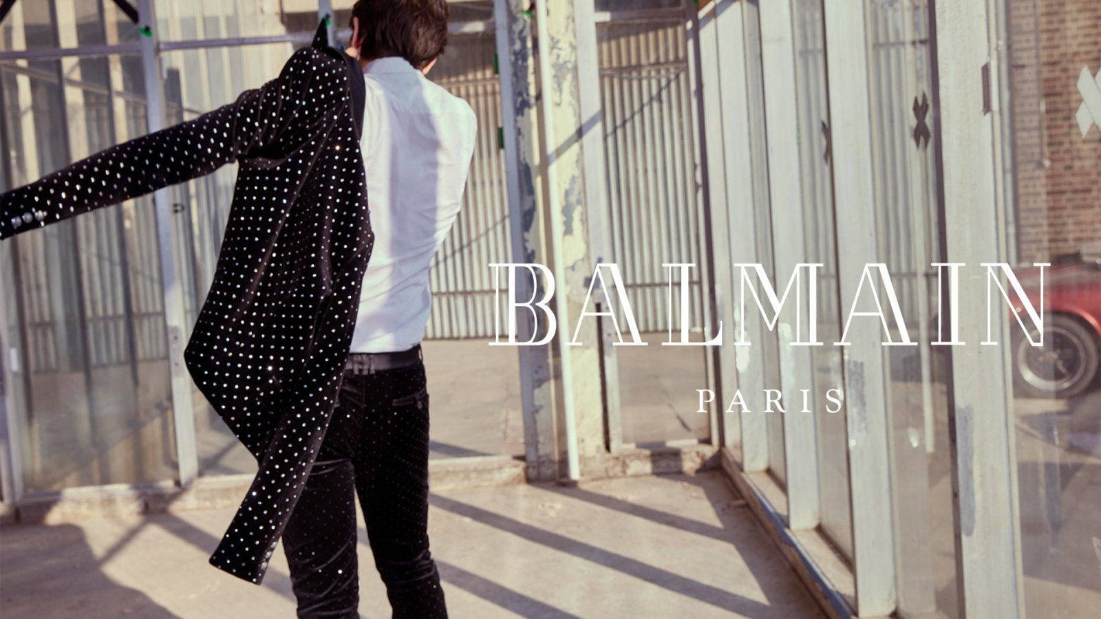 身穿圓點西裝外套的男子在陽光明媚的城市小巷中走遠，帶有Balmain Paris文字。