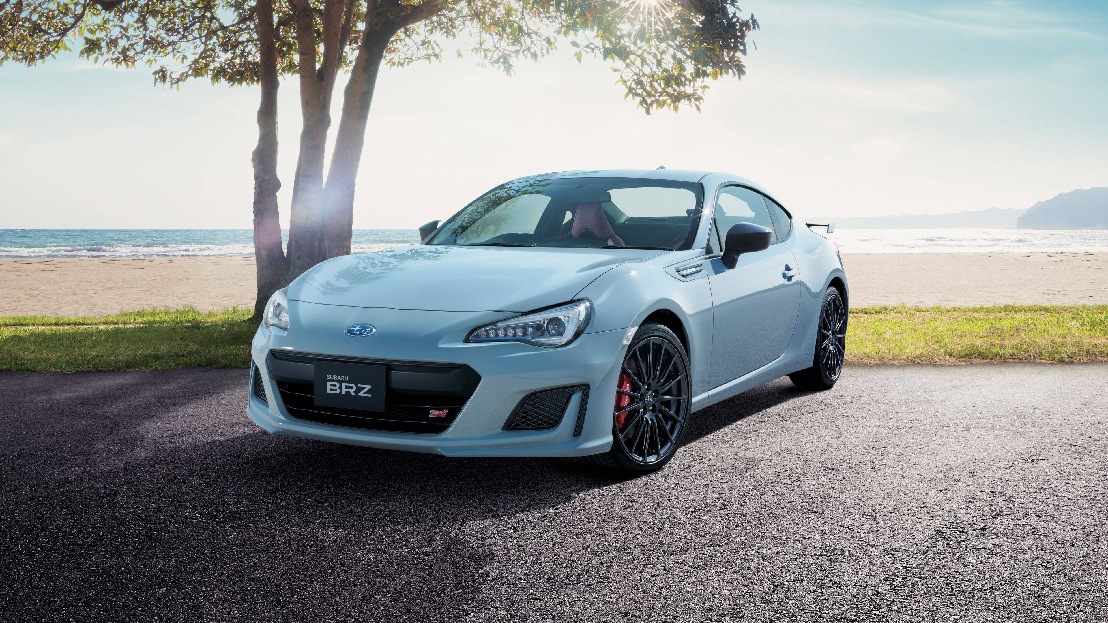 斯巴魯BRZ海灘停泊 壁紙 — 一架時尚的淺藍色Subaru BRZ跑車停泊在鋪砌小徑上，旁邊有樹木，背景是海洋和沙灘，在明亮的陽光下。
