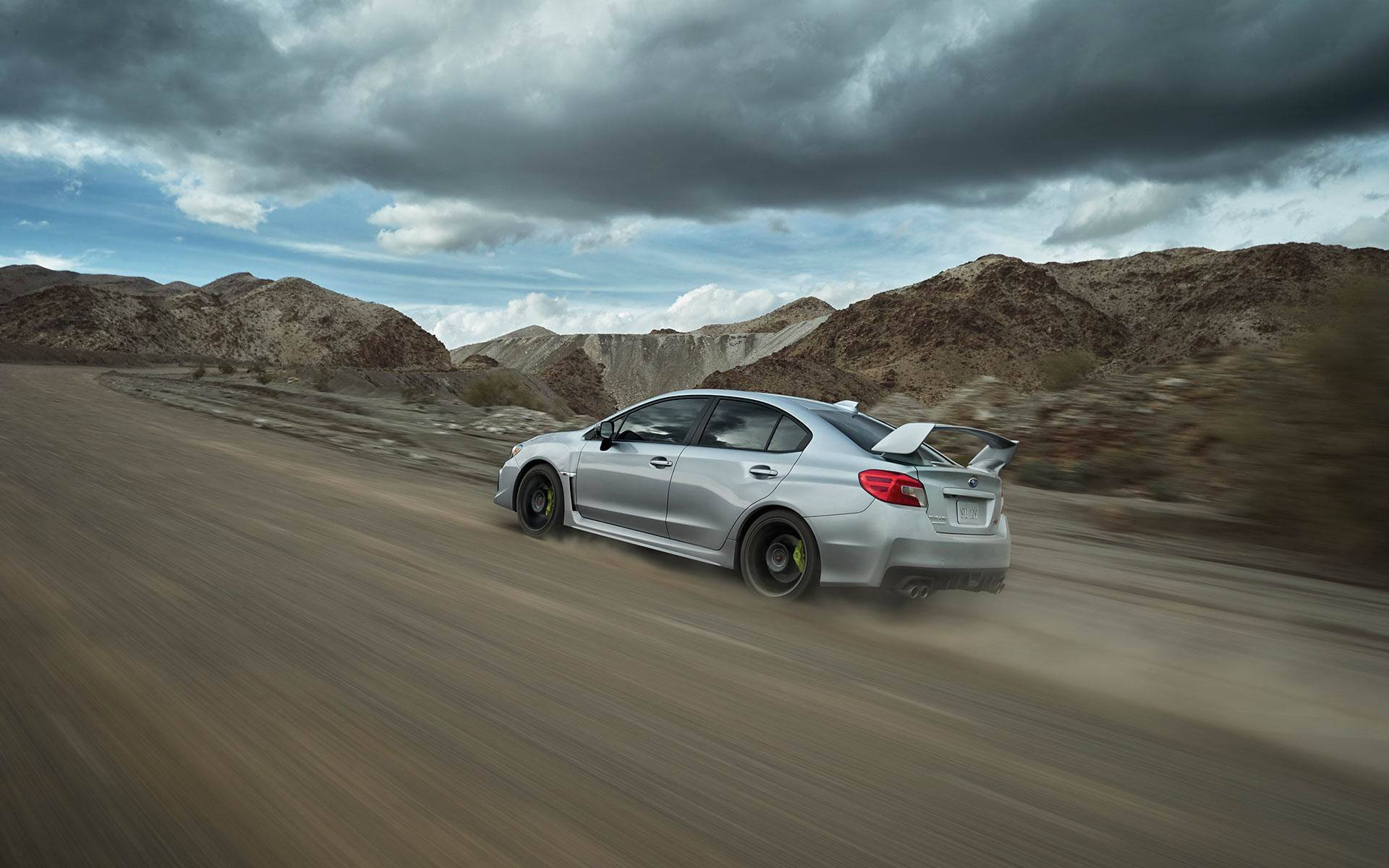 斯巴魯Impreza高速衝坡 壁紙 — 一輛銀色Subaru WRX STI在戲劇性的多雲天空下，疾速穿越塵土飛揚的沙漠峽谷。