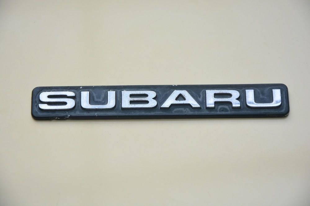 斯巴魯標誌名稱徽章 壁紙 — 銀色 Subaru 標誌喺米色車頭蓋上嘅特寫，光線柔和，表面有輕微磨損。