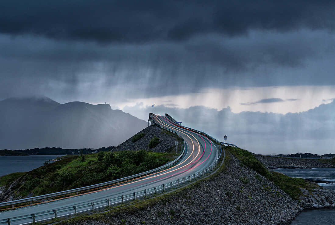 在風暴天空下，一條蜿蜒的道路越過山丘，帶有汽車燈光的條紋。