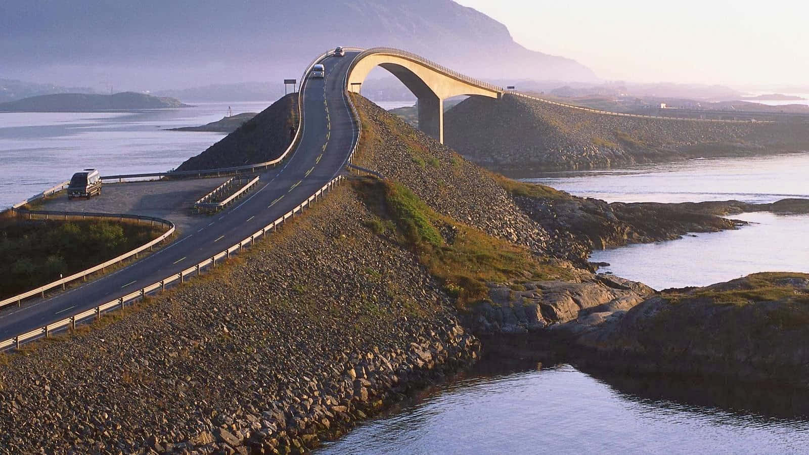 一座彎曲的現代橋樑橫跨岩石峽灣，汽車在日落時分行駛。