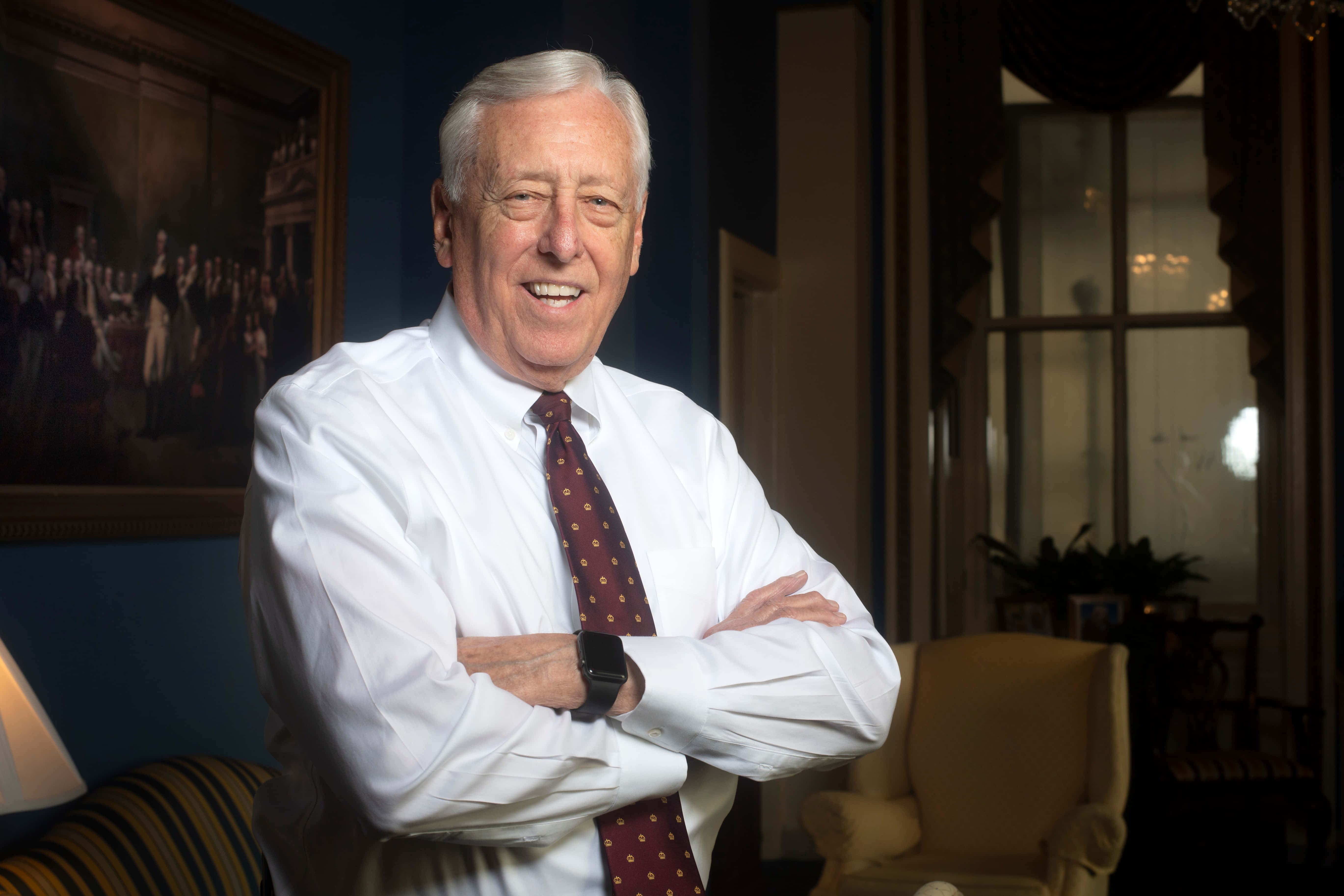 斯泰尼·霍耶微笑雙臂交疊 壁紙 — 一位白髮年長男性在裝飾華麗的房間裡，藍色牆壁和裱框藝術品襯托下，自信地交叉雙臂微笑。