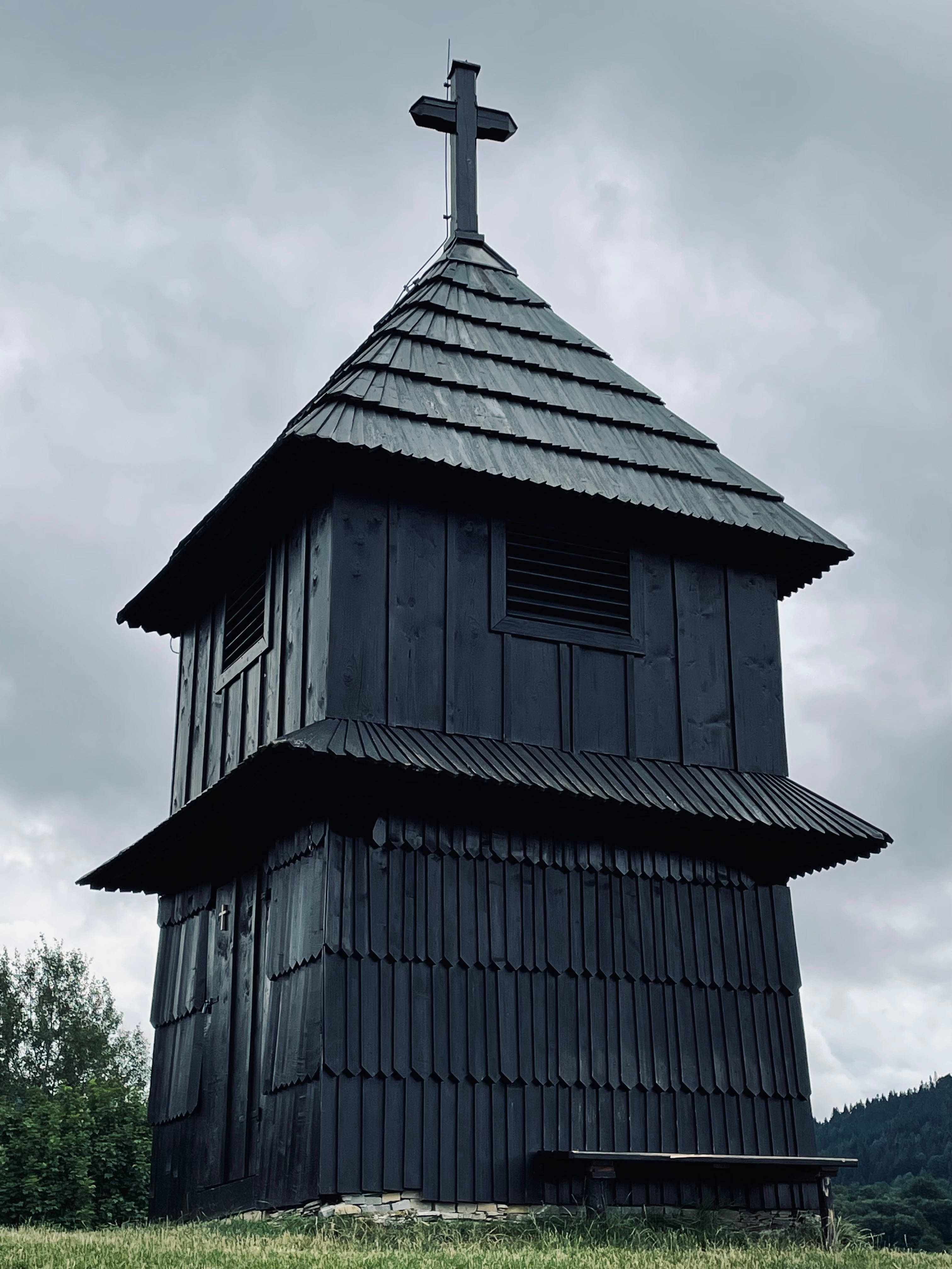 斯洛伐克 Zvonica u Marunov 歷史地標 壁紙 — 一個深色木製教堂鐘樓帶有十字架，在陰天下的草地上莊嚴地矗立。