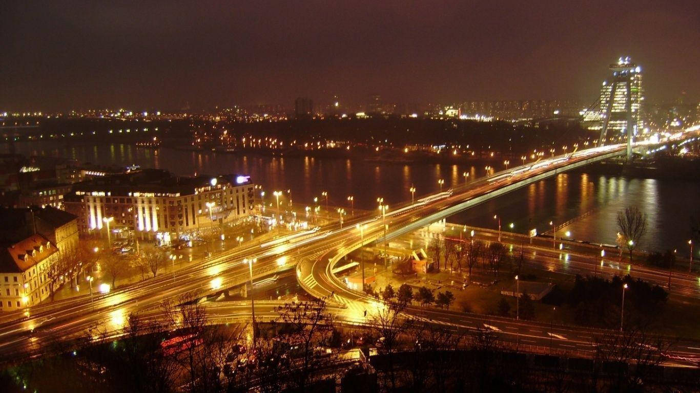 斯洛伐克城市夜景道路 壁紙 — 充滿活力的夜景城市景觀，發光的燈光倒映在河上，車流的光跡流過橋樑。