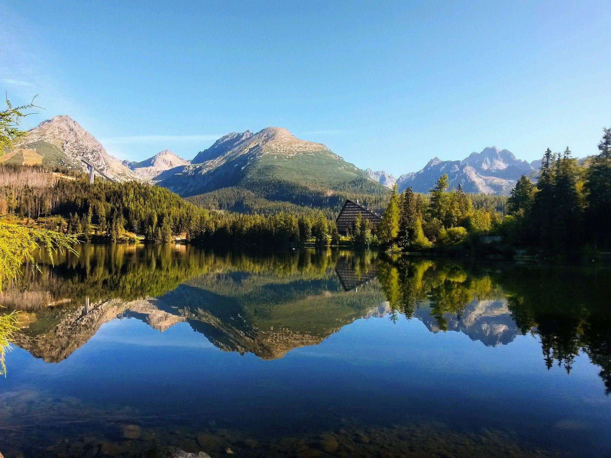 斯洛伐克山湖倒影 壁紙 — 寧靜的山湖映照著鋸齒狀的山峰和一座木屋，天空清澈湛藍。