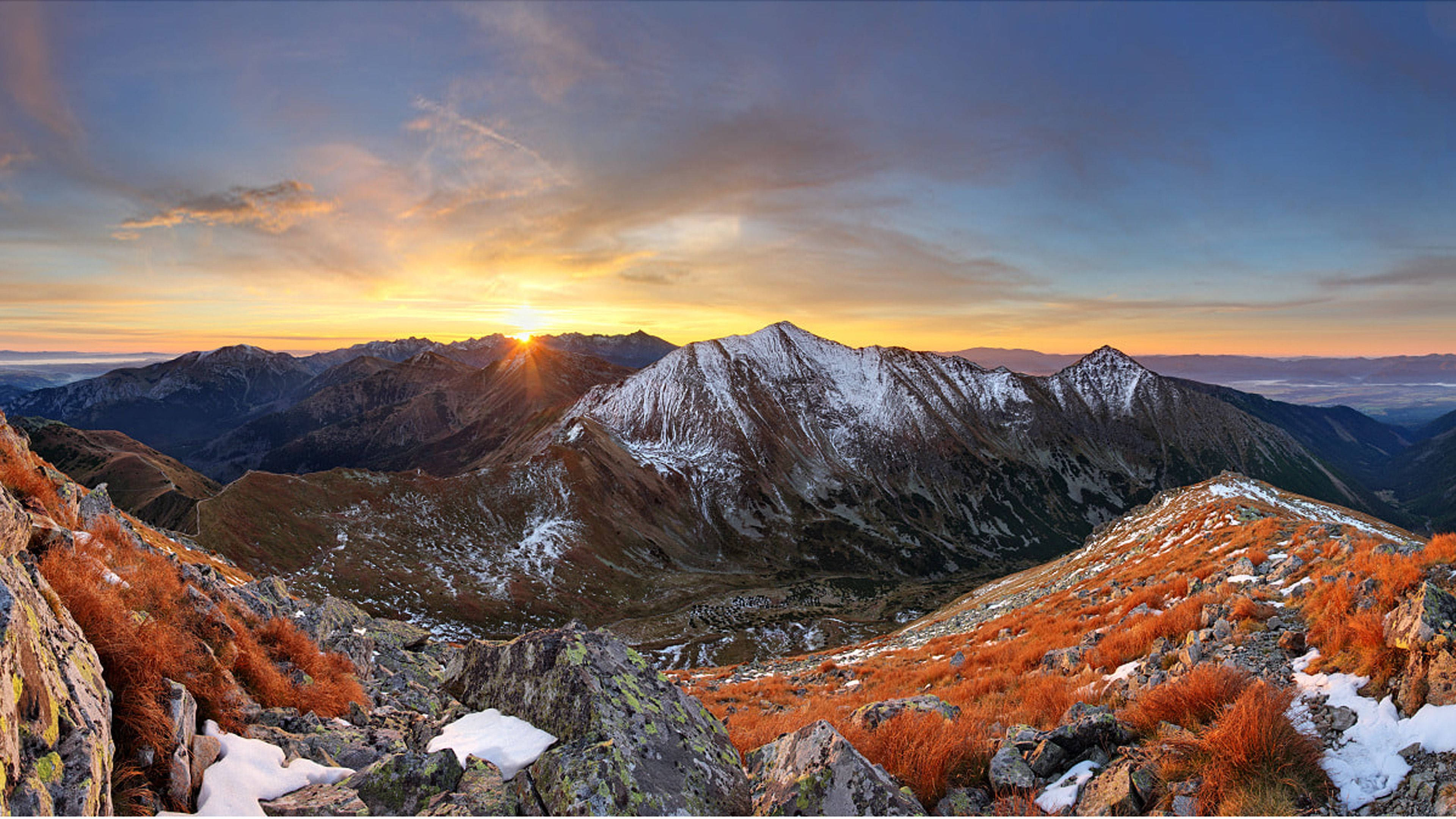 斯洛伐克群山日落 壁紙 — 夕陽照耀在積雪的山峰上，前景是充滿活力的橙色草叢。