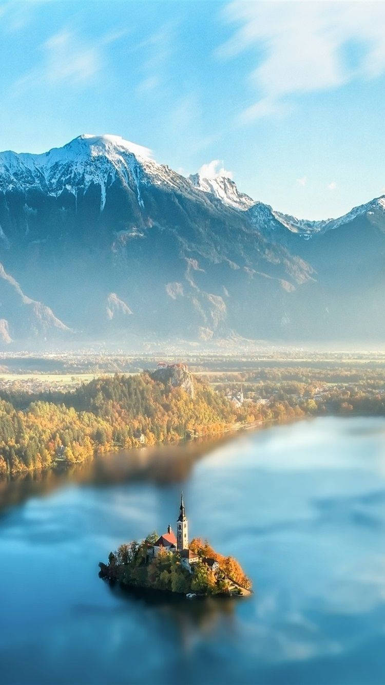 斯洛維尼亞湖畔的晴空美景 壁紙 — 一個寧靜的湖泊，湖中小島上有一座教堂，周圍環繞著秋天的樹木和白雪覆蓋的山脈，天空湛藍清澈。
