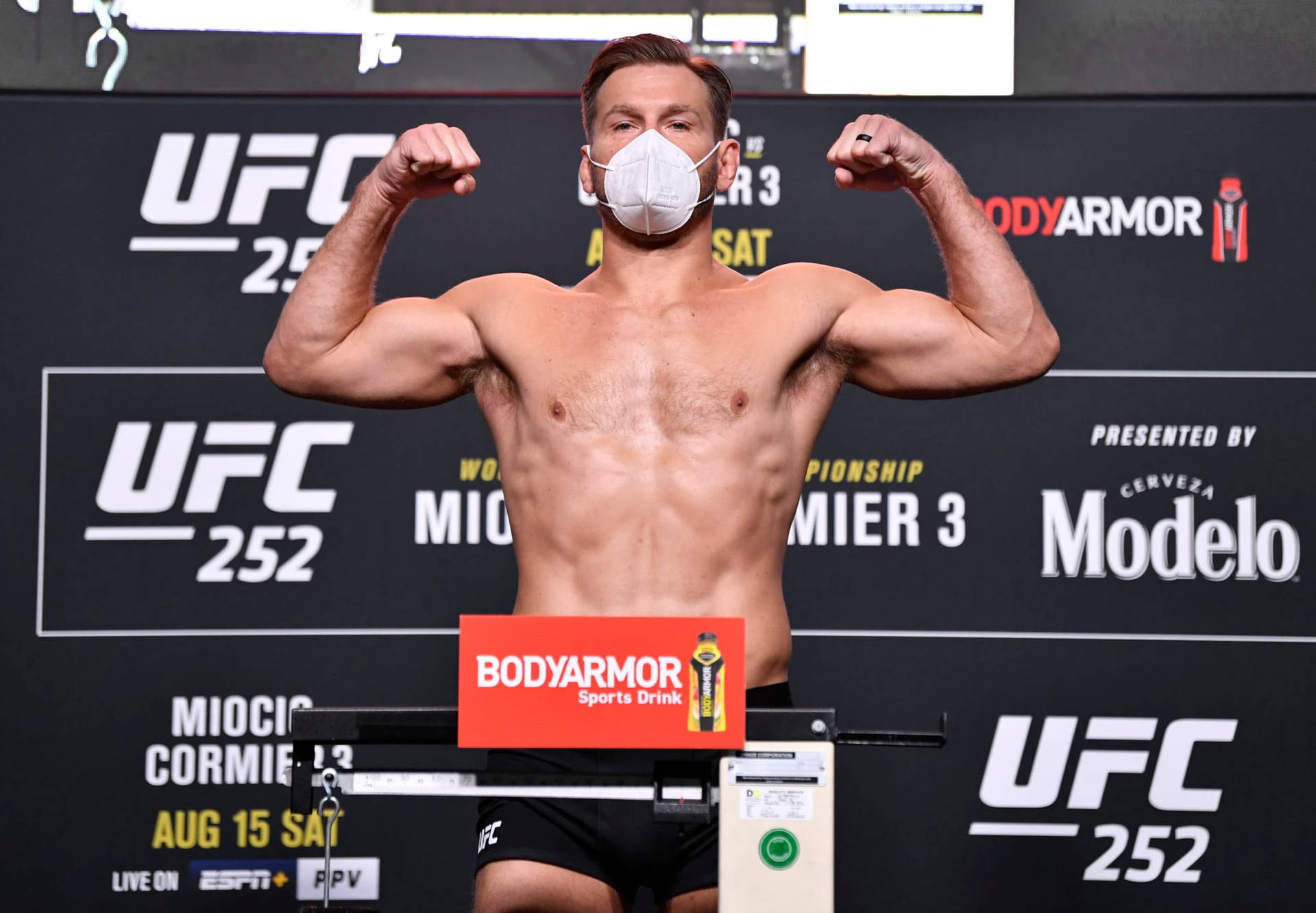 斯蒂普·米奧奇UFC 252稱重儀式 壁紙 — 一位肌肉發達的UFC拳手在稱重活動中戴著口罩，身後有贊助商標誌，彎曲手臂展示肌肉。