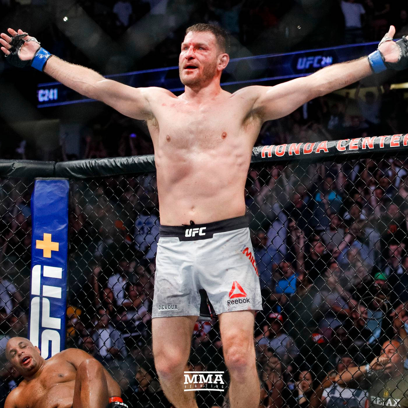 斯蒂普·米奧奇克與落敗對手 壁紙 — 一位勝利的MMA拳手在八角籠中高舉雙臂，慶祝勝利，而他的對手則在明亮的場館燈光下倒地敗北。