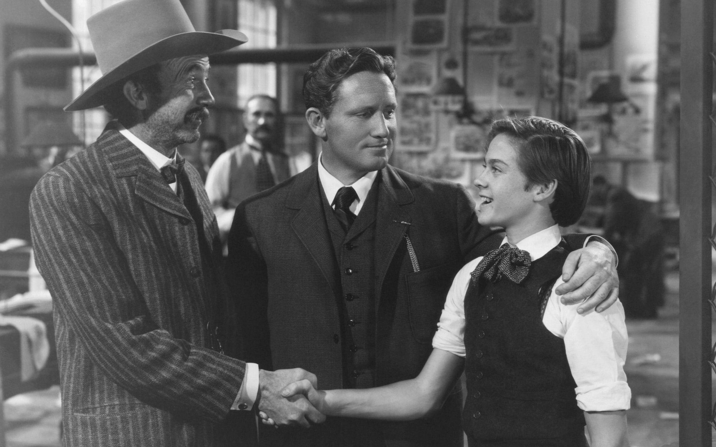 斯賓塞·崔西主演1939年電影《史丹利與利文斯通》 壁紙 — 三位身穿復古西裝和帽子的男士在一個堆滿海報的雜亂工作室裏熱情握手。