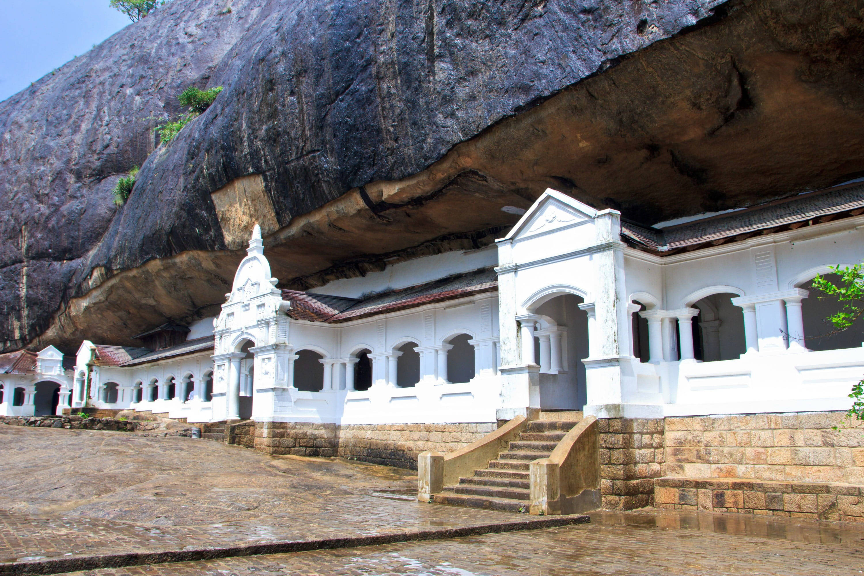 斯里蘭卡丹布拉洞穴寺 壁紙 — 白色殖民風格寺廟建築群，坐落於巨大深色岩石懸垂下方，地面濕滑。