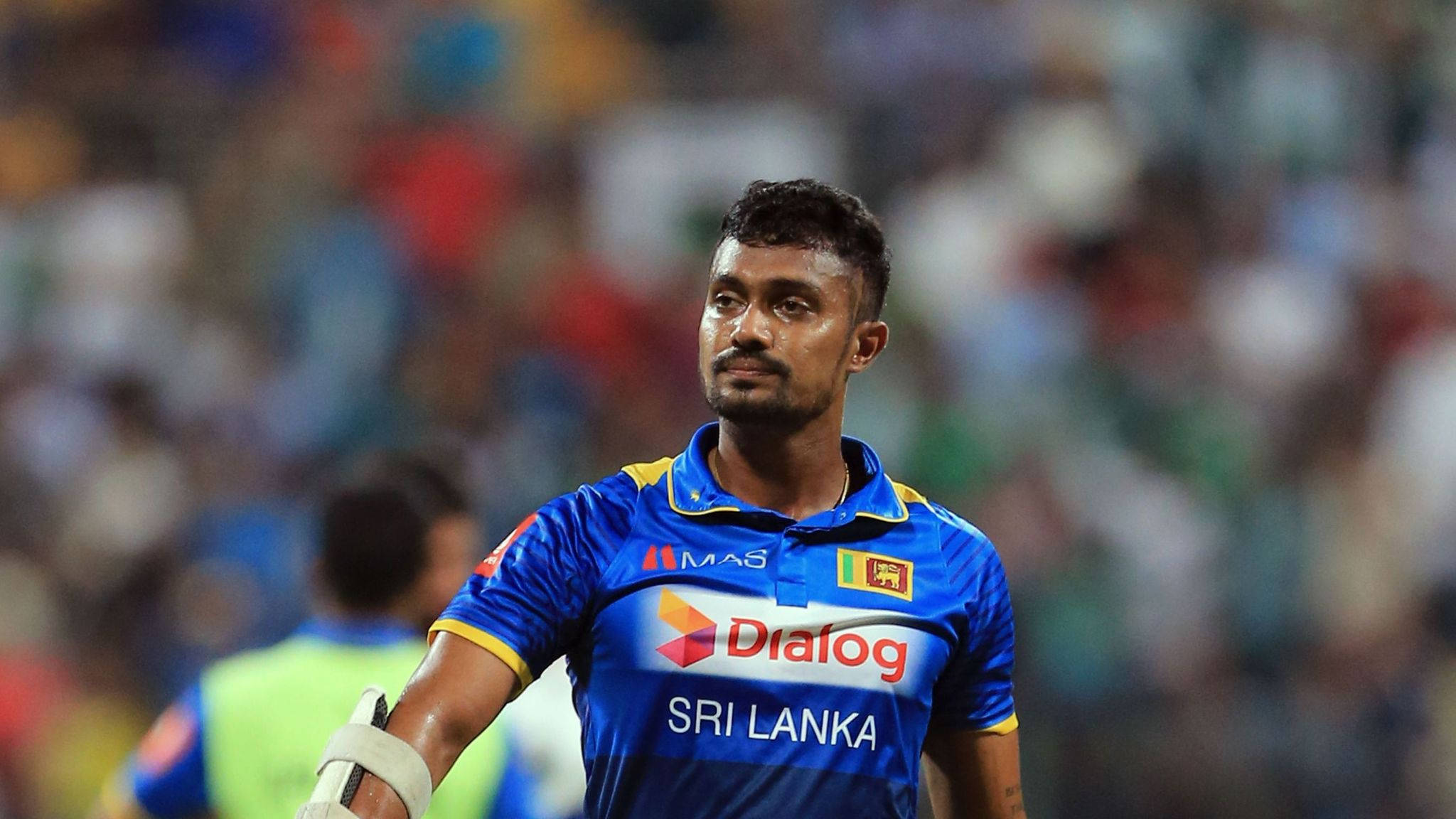斯里蘭卡板球運動員Dhanushka Gunathilaka 壁紙 — 一名身穿藍色球衣的斯里蘭卡板球運動員專注地站在球場上，背景是模糊的體育場人群。