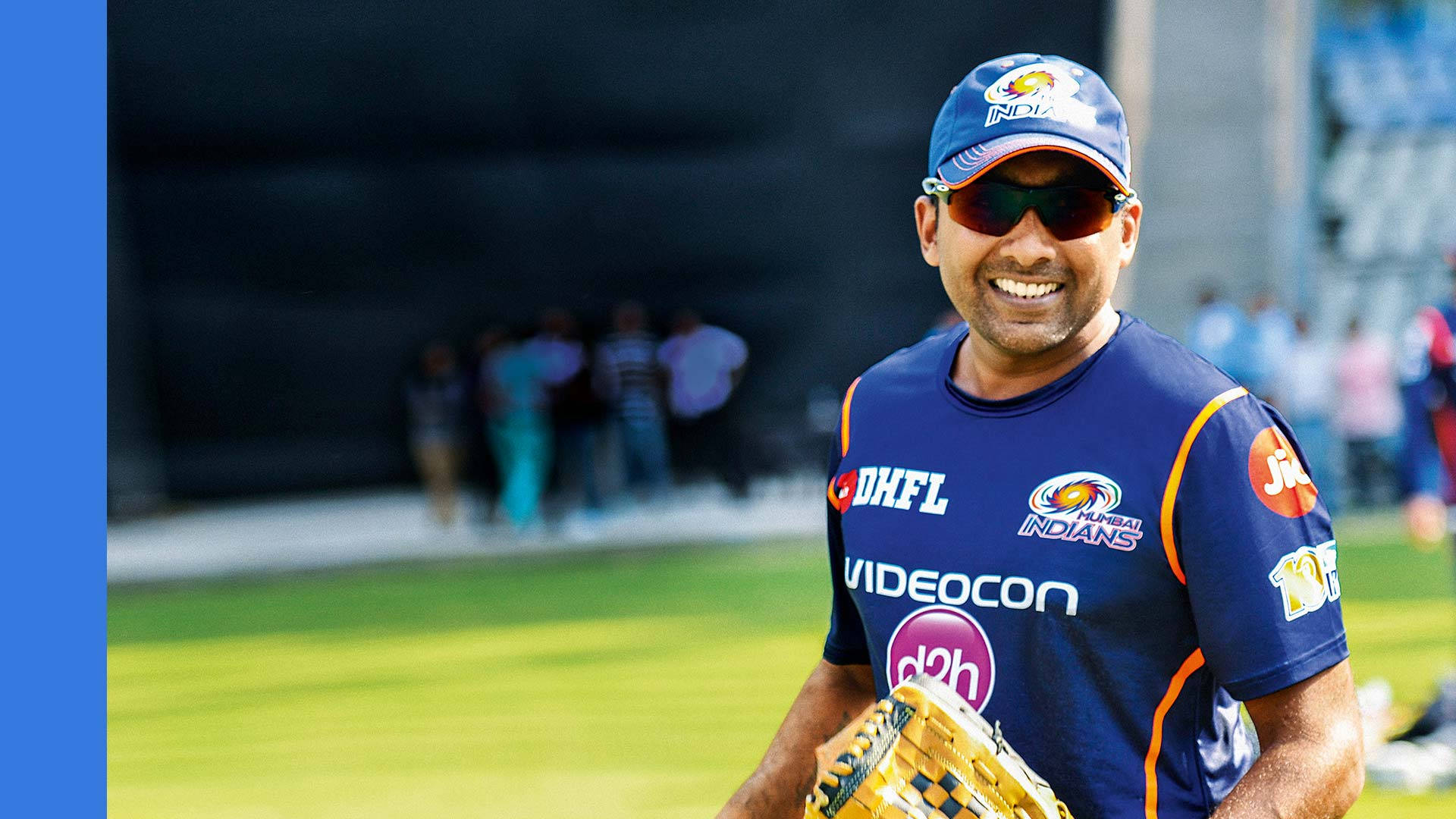 斯里蘭卡板球運動員Mahela Jayawardene 壁紙 — 一位微笑的板球運動員身穿藍色Mumbai Indians球衣和帽子，在陽光明媚的球場上拿著黃色手套。