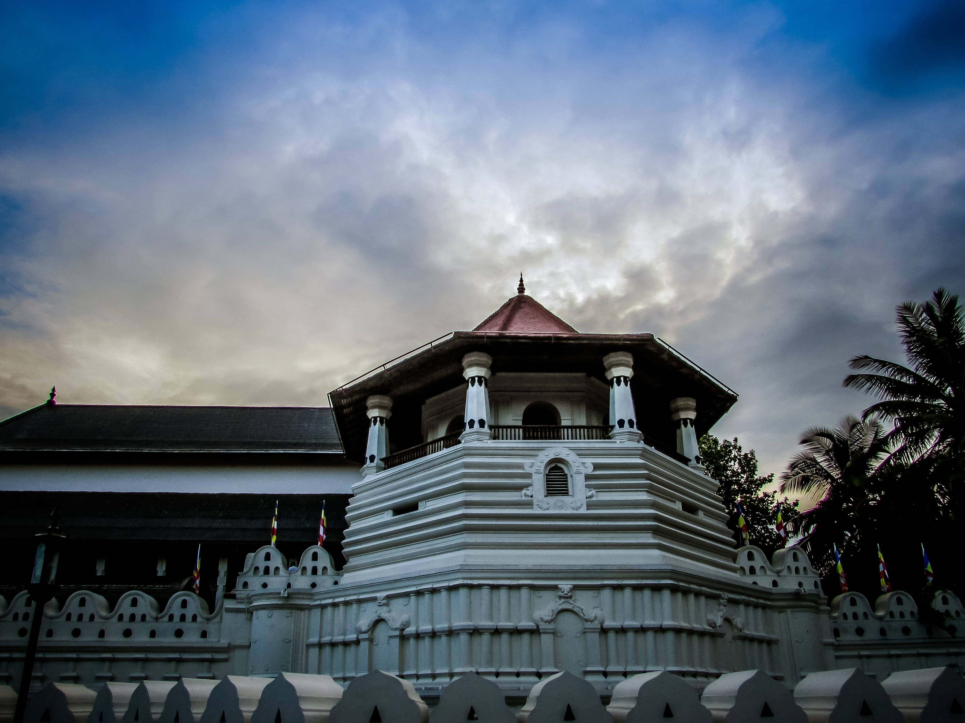 斯里蘭卡聖牙舍利寺 壁紙 — 一座白色分層寺廟，紅色屋頂，矗立在戲劇性的多雲天空下，附近有棕櫚樹。