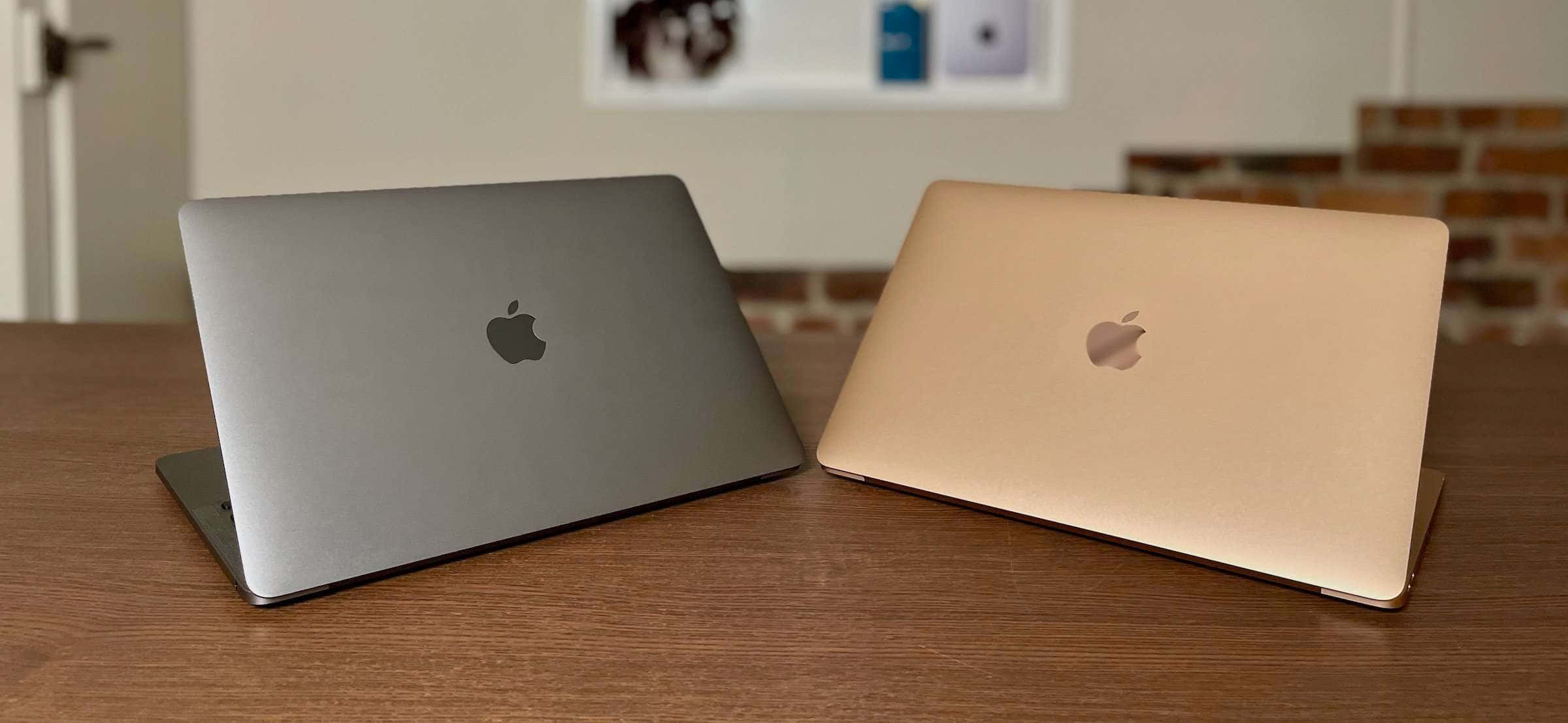 新一代計算——MacBook 壁紙 — 兩部 Apple 手提電腦銀色和金色合上放在木桌上，背景有磚牆和裱框藝術品。