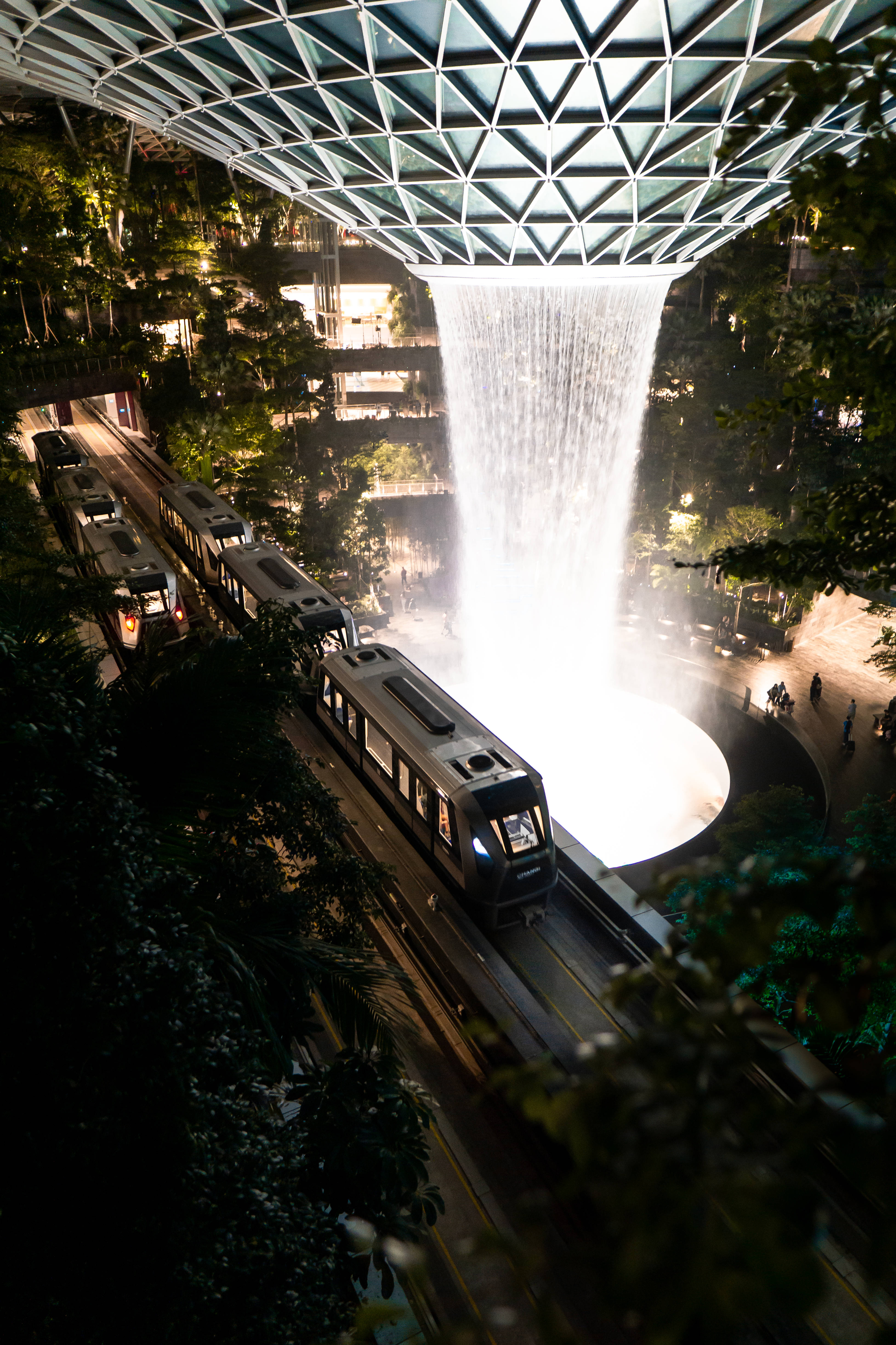新加坡 Jewel Changi Airport HSBC 雨旋風 iPhone 藝術壁紙 壁紙 — 夜晚，一列火車在幾何玻璃天花下，於一個大型室內瀑布下方滑行而過。