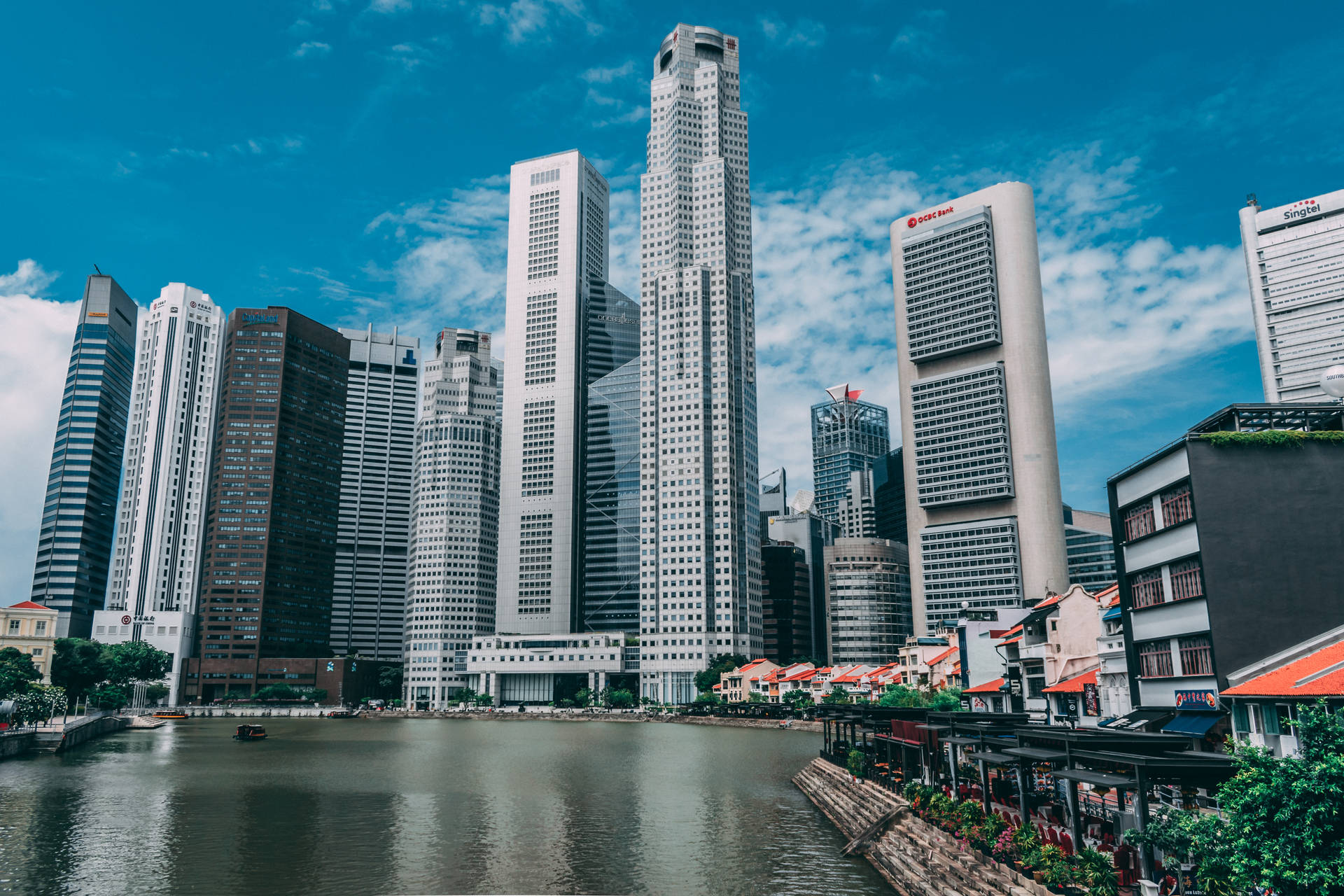 高聳的城市建築倒映在河中，旁邊是色彩繽紛的店屋，天空晴朗。