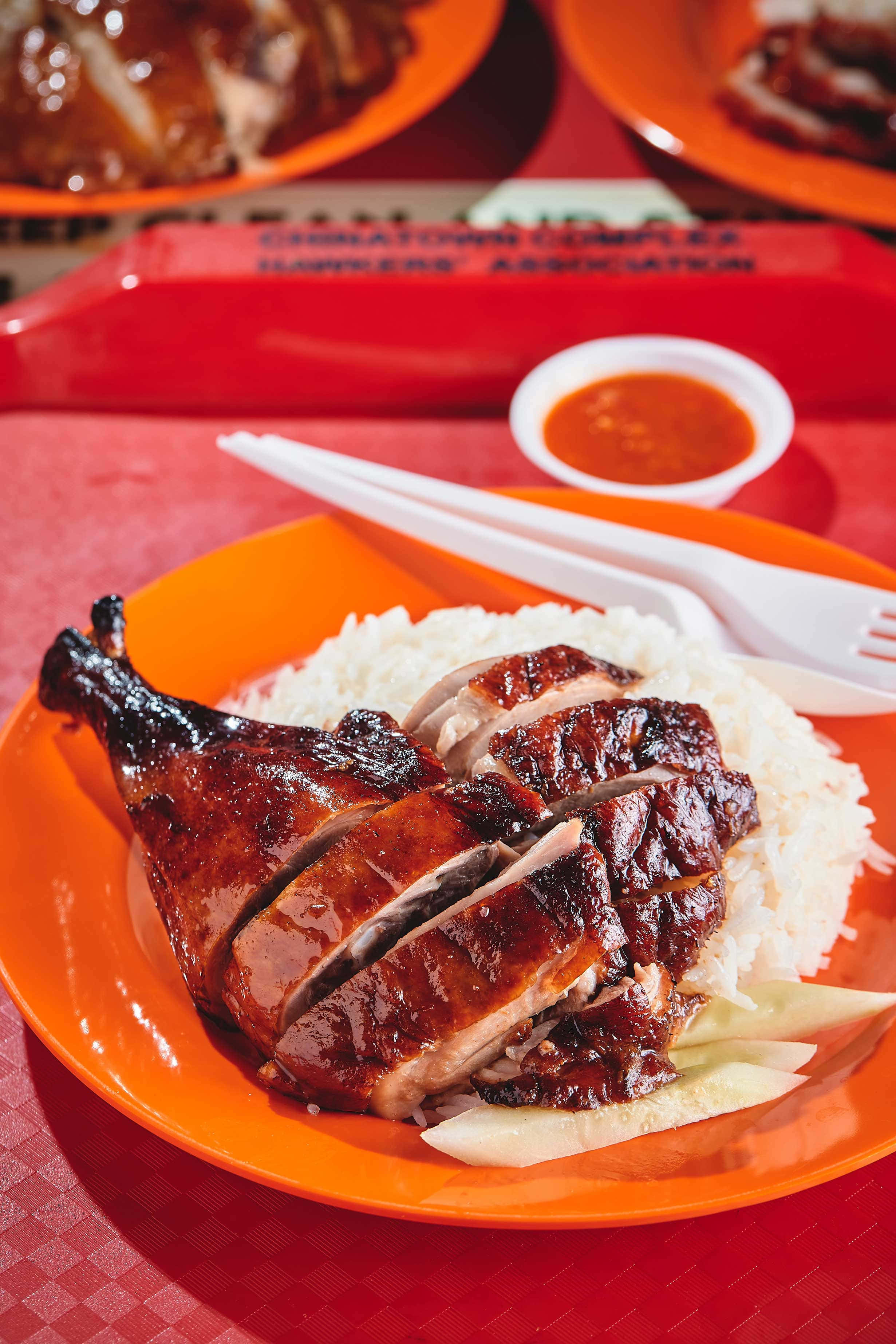 新加坡牛車水美食中心鴨肉飯 壁紙 — 閃閃發光的烤鴨，焦糖化的皮放在白飯上，放在一個充滿活力的橙色碟子上，配上辣椒醬和筷子。