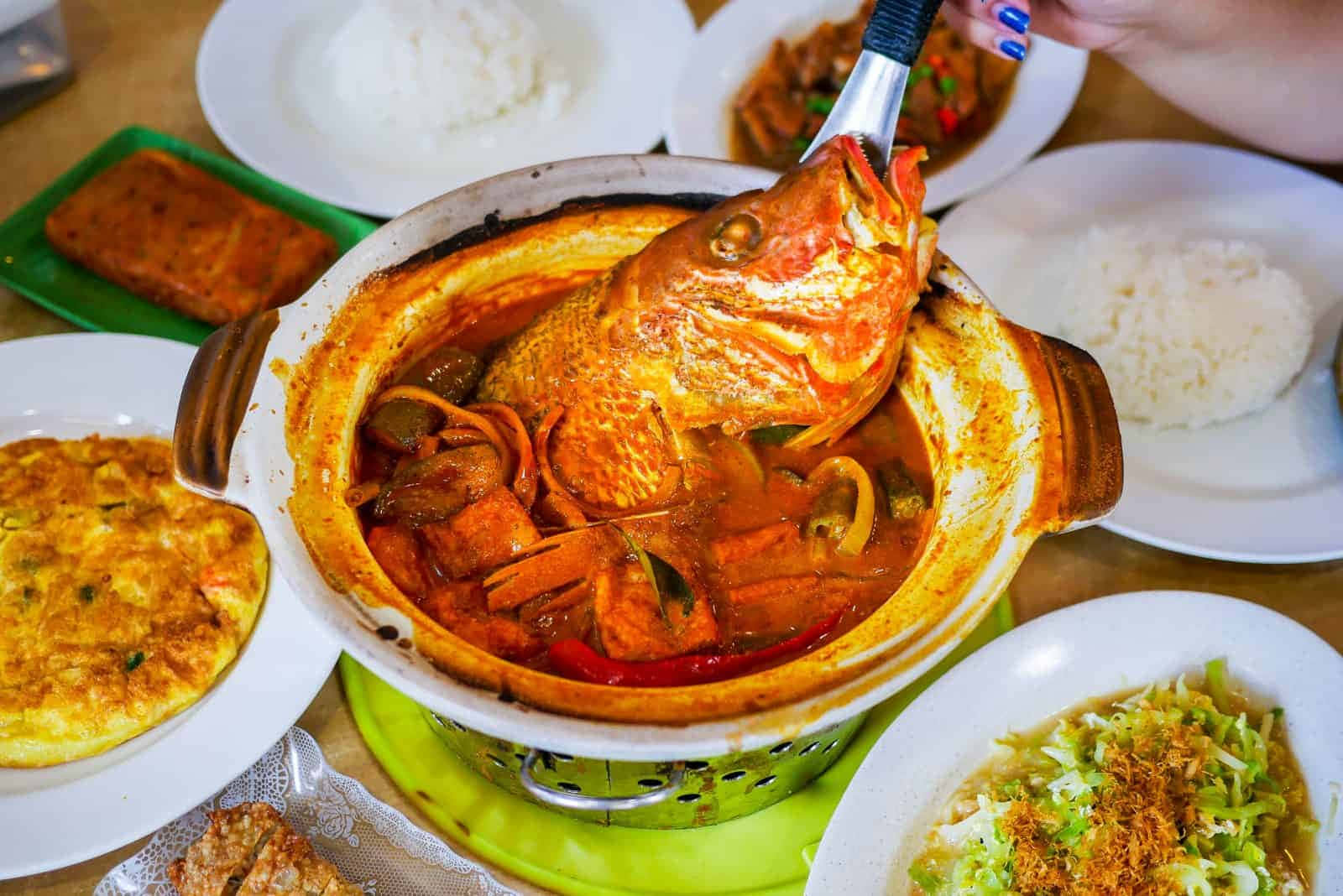 新加坡特色美食魚頭咖喱 壁紙 — 一個冒煙的陶鍋，裡面裝著辛辣的魚咖喱，用鉗子夾起整條魚，周圍是米飯和配菜。