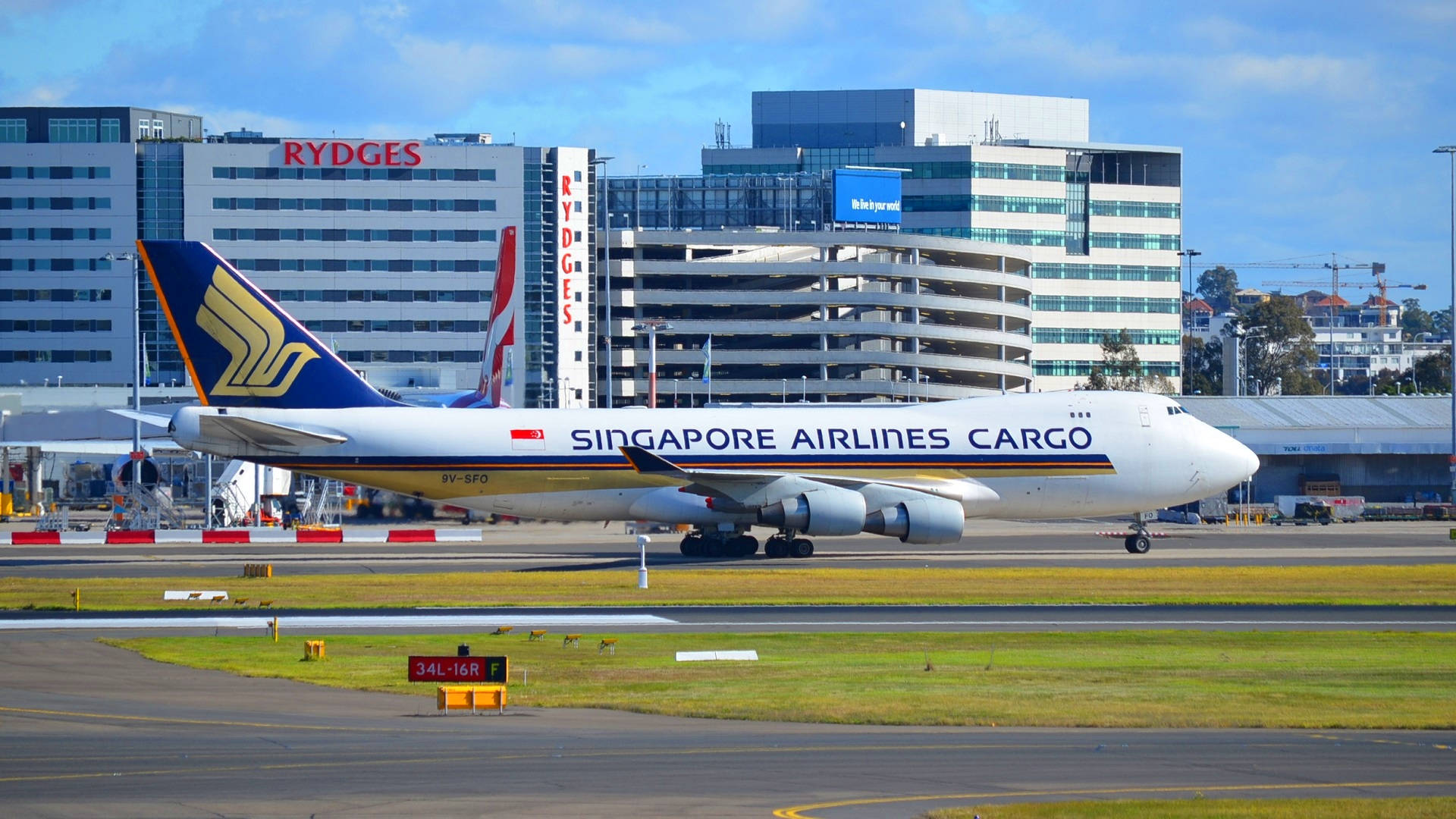 新加坡航空機場貨機 壁紙 — 一架新加坡航空貨運波音747在陽光明媚的機場停機坪上滑行，背景有現代建築和清澈的藍天。