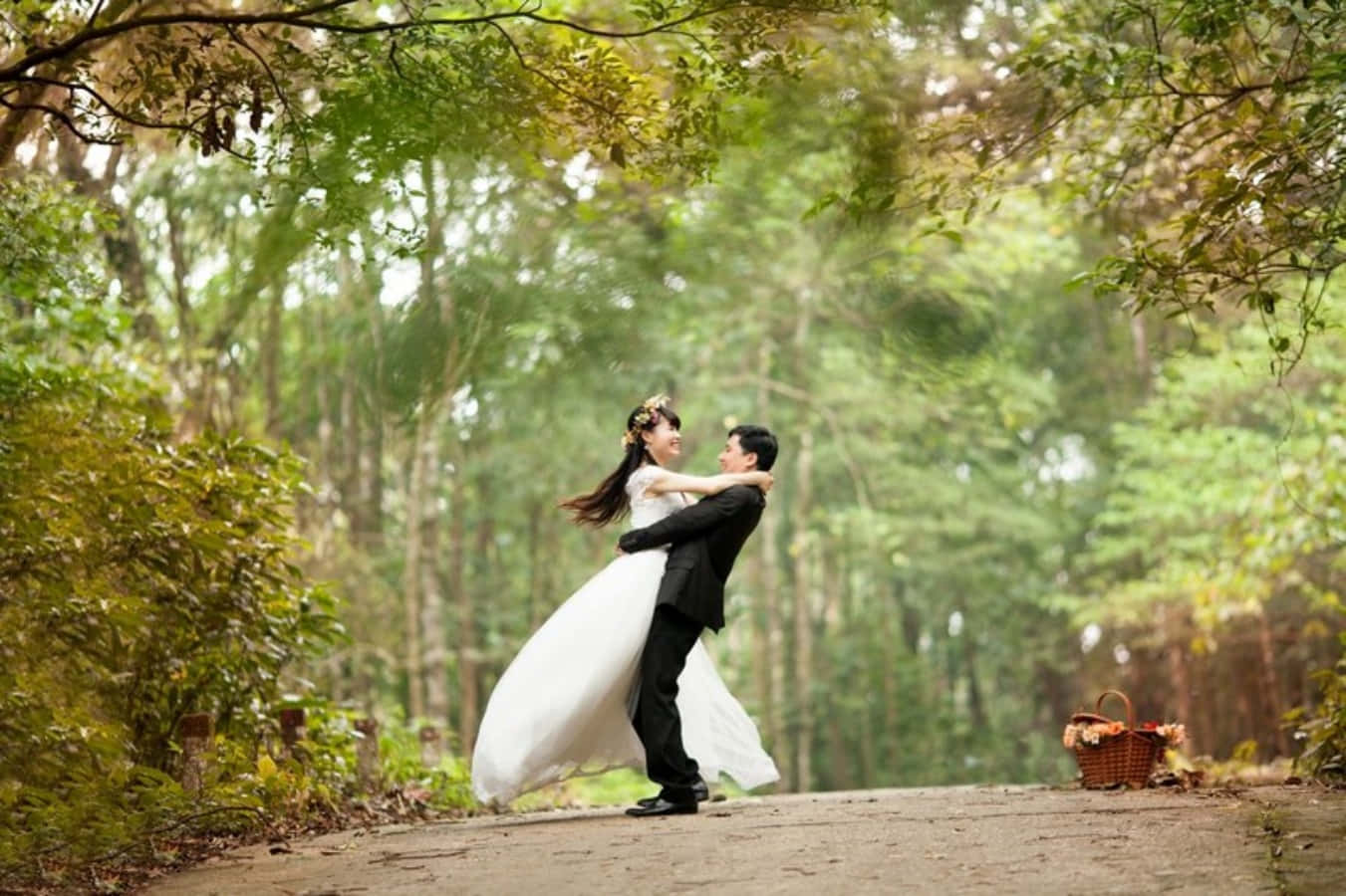 Wedding Couple 桌布 壁紙 — 新娘和新郎在森林小徑上快樂地擁抱旋轉，陽光穿過綠色樹冠灑落下來。