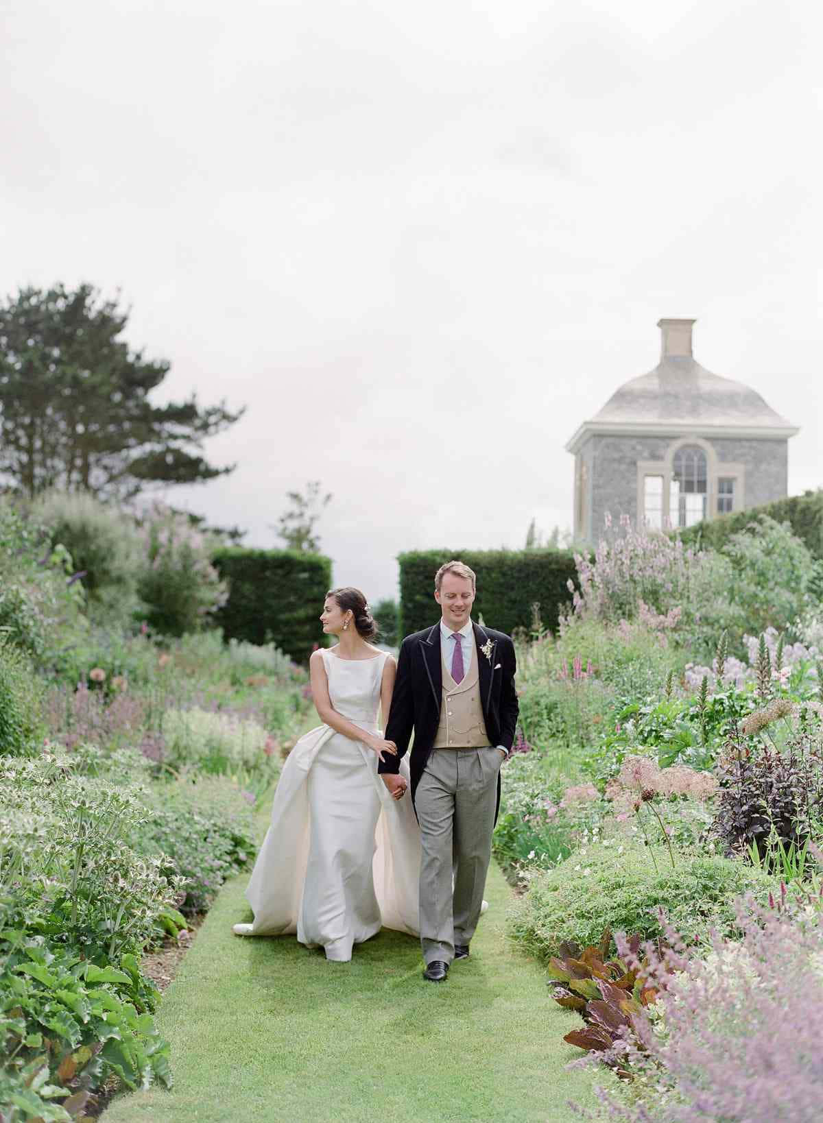 Garden Wedding 桌布 壁紙 — 新娘和新郎手拖手，沿著一條草地小徑穿過一個茂盛的花園，園內有盛開的花朵和遠處的一座石頭涼亭。