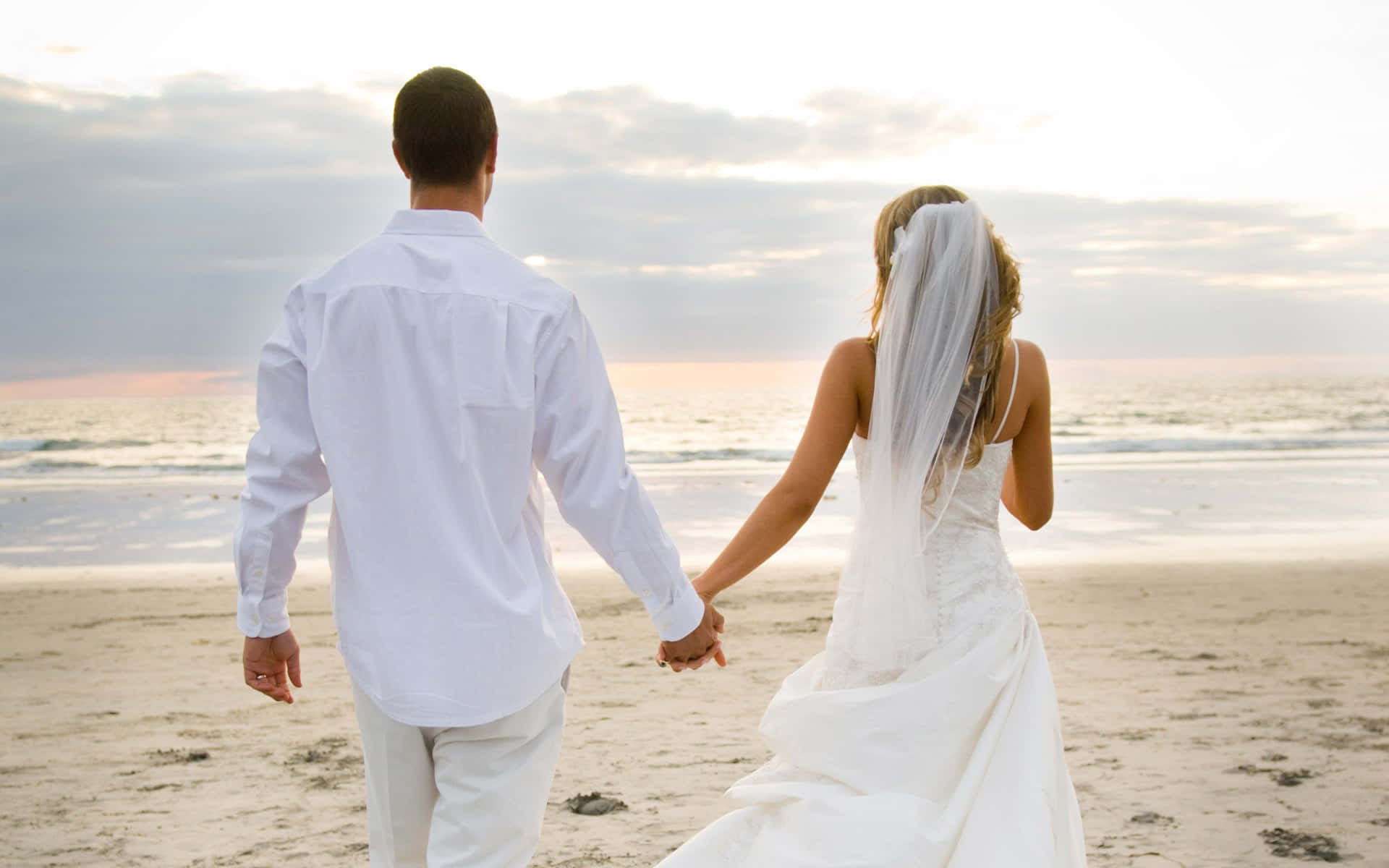 Wedding Couple 桌布 壁紙 — 新娘和新郎手牽手沿著沙灘漫步，夕陽西下，溫柔的海浪和柔和的金色光芒。