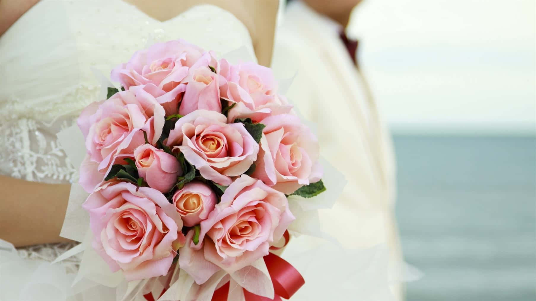 Wedding Bouquet 桌布 壁紙 — 新娘手持一束豐盛的粉紅玫瑰，繫著紅色絲帶，背景是模糊的海洋。