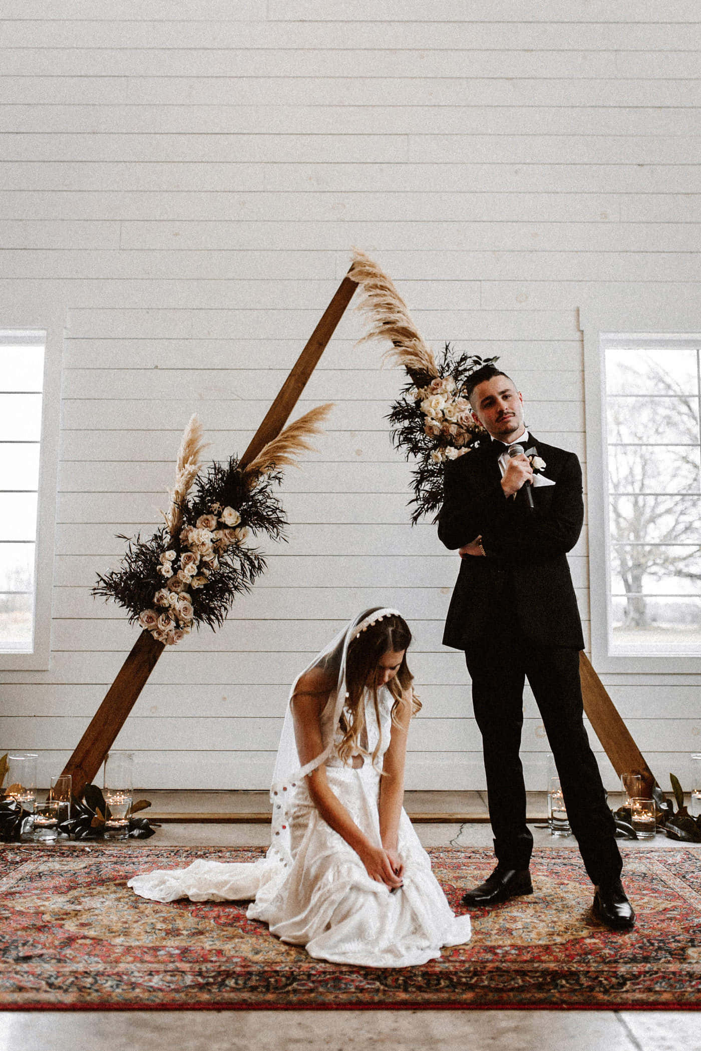 Black And White Wedding 桌布 壁紙 — 新娘跪在圖案地毯上，下方是裝飾著乾草和鮮花的鄉村風格木拱門，新郎身穿黑色西裝站在她旁邊。