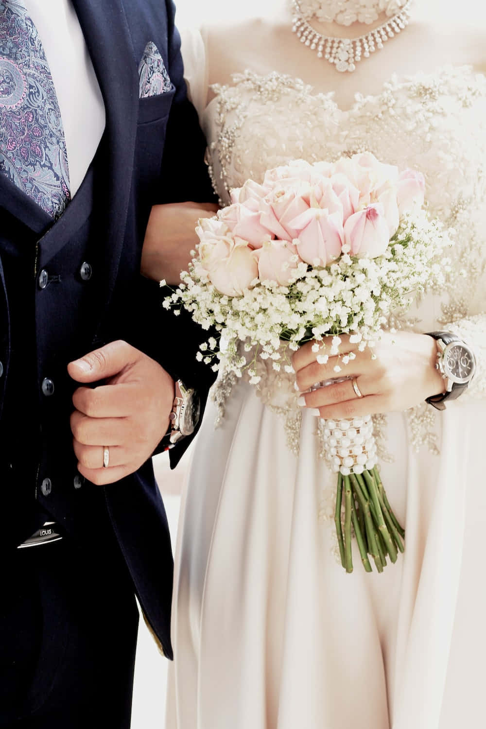新婚夫婦喜慶擁抱，共享幸福時刻 壁紙 — 身穿優雅婚禮服飾的新娘和新郎手牽手，新娘緊握一束粉紅玫瑰和滿天星花球。