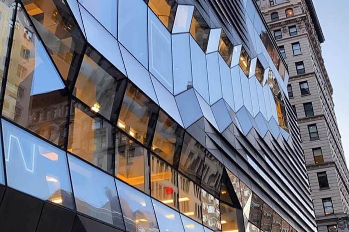 新學校的璀璨窗戶 壁紙 — 一座現代玻璃建築，幾何形切面反射溫暖的室內燈光及相鄰的歷史建築，背景是晴朗的天空。